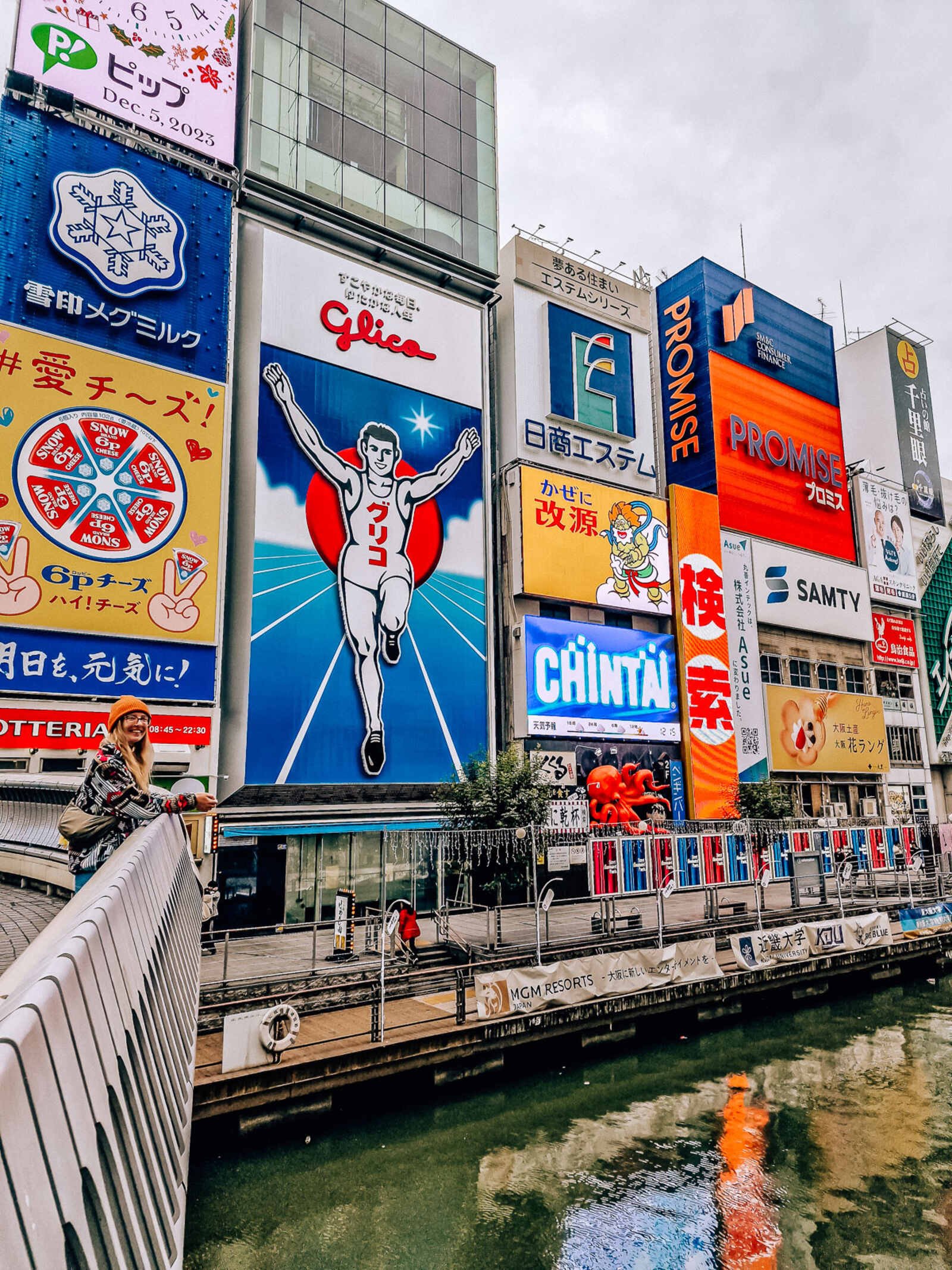 Daytime with lots of colourful adverts and billboards on the side of buildings along a river including a large portrait of a running man with his arms in the air - this is the osaka glico running man sign