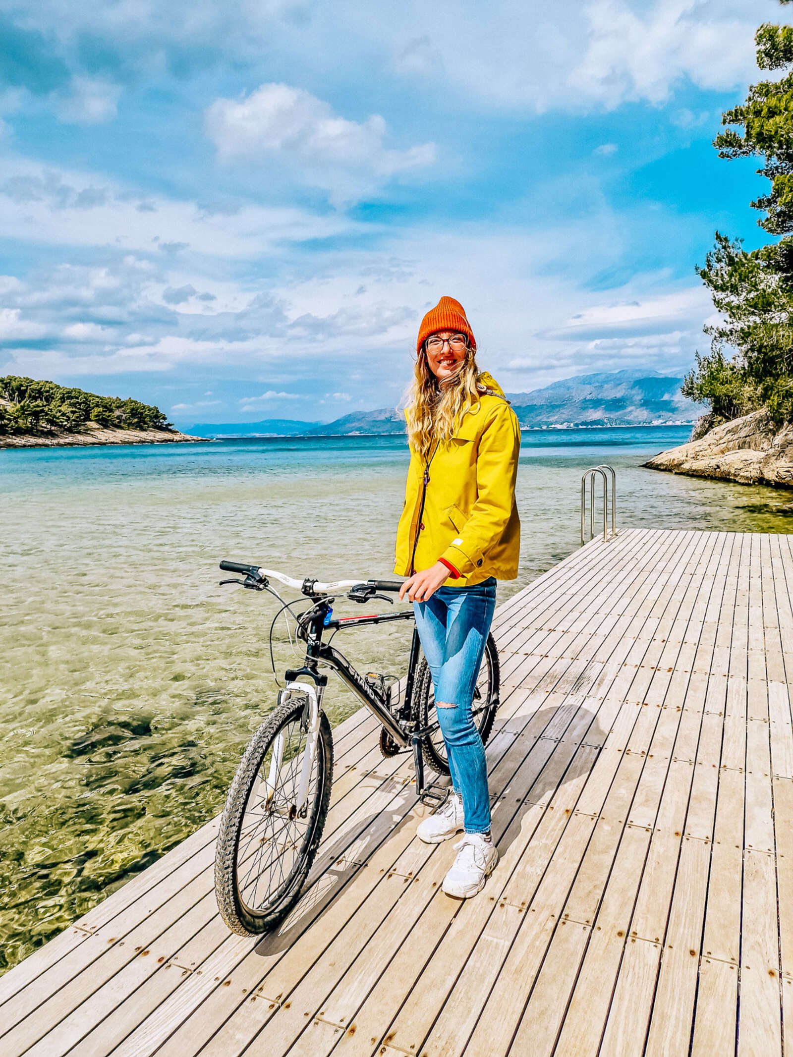 girl in yellow jacket, holding a bike on a dock next to clear blue sea in a bay
