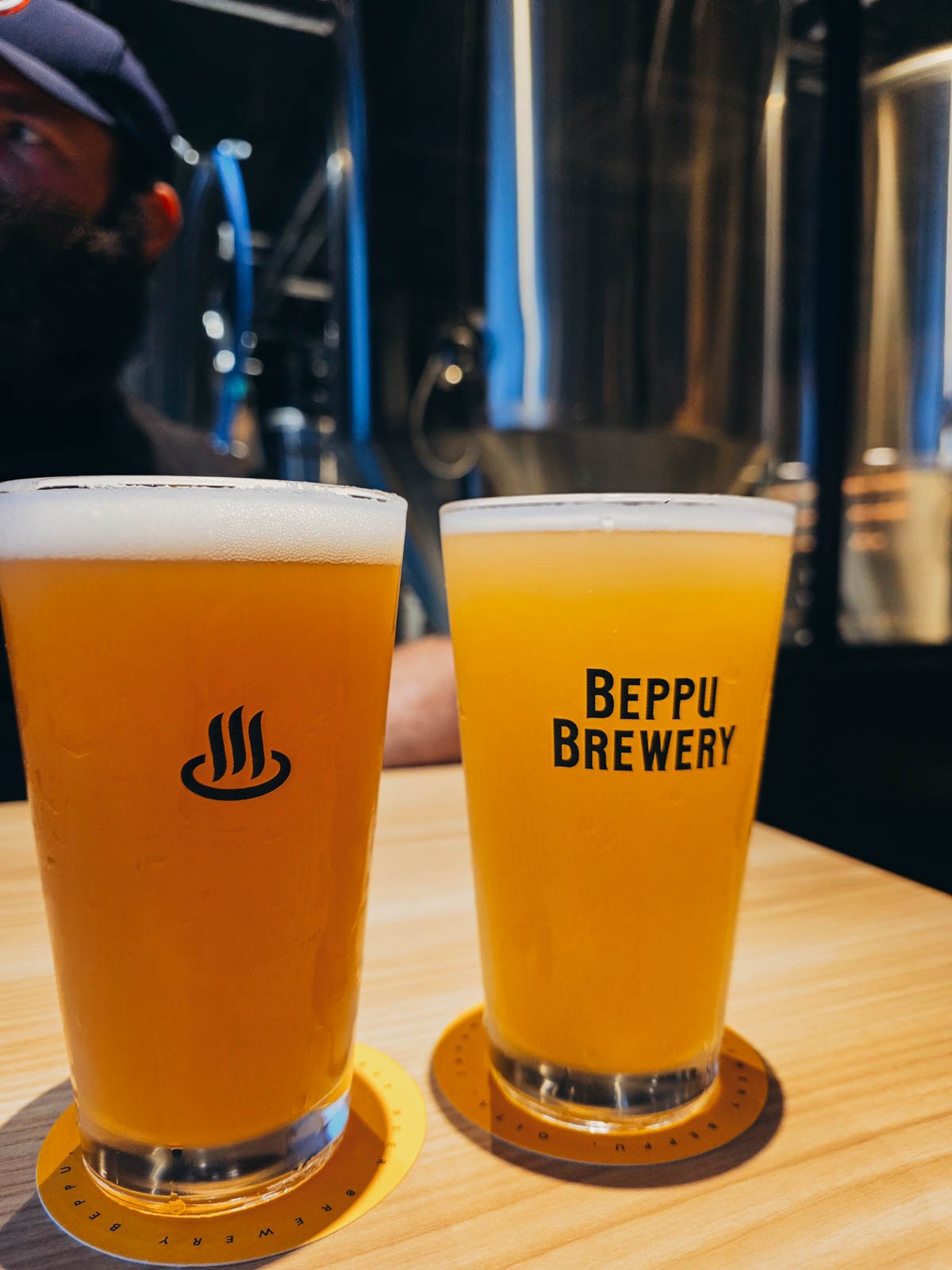 Two full glasses of craft beer on a wooden table at a brewery, one of the glasses has "Beppu brewery" printed on it