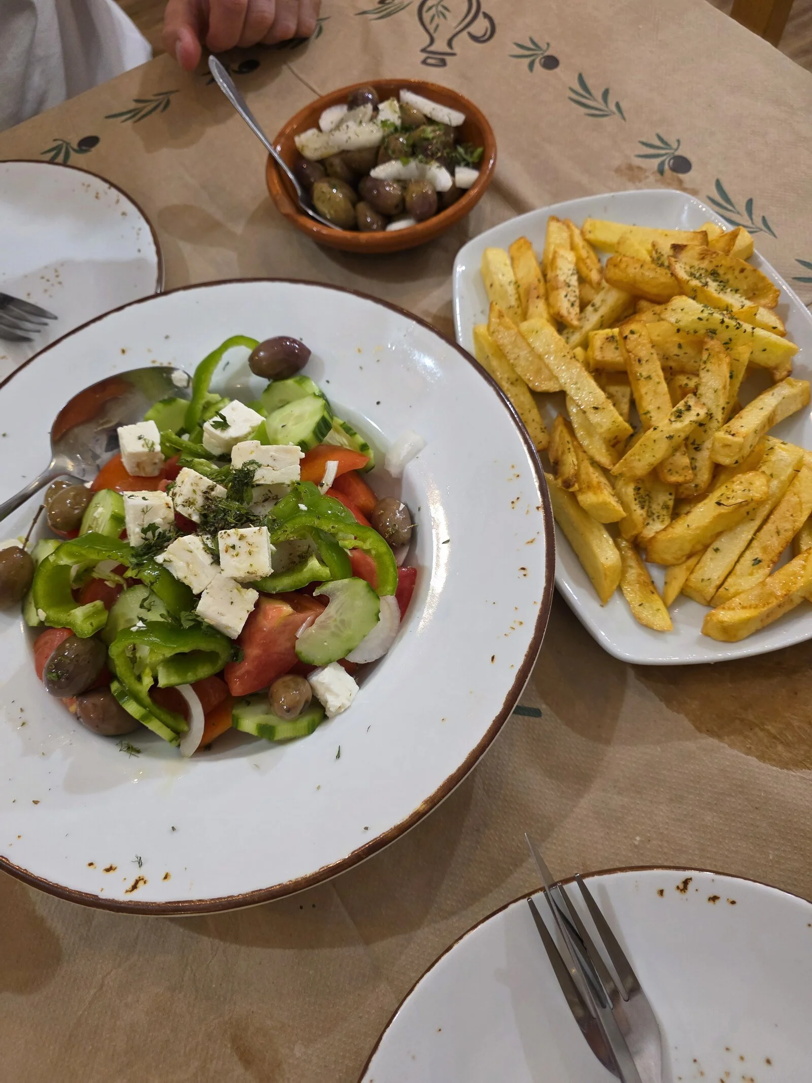 A Greek style salad, fries, and a bowl of olives in white dishes and a brown table.