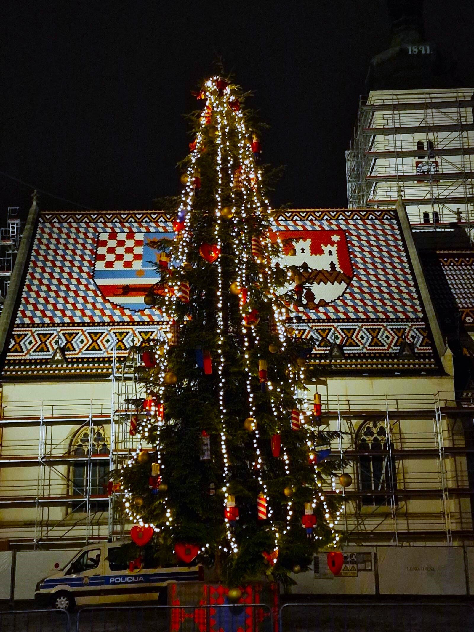 a large christmas tree with red decorations and white lights stands in front of a church with a red and white checked rooftop showing two coats of arms. Picture is taken at night
