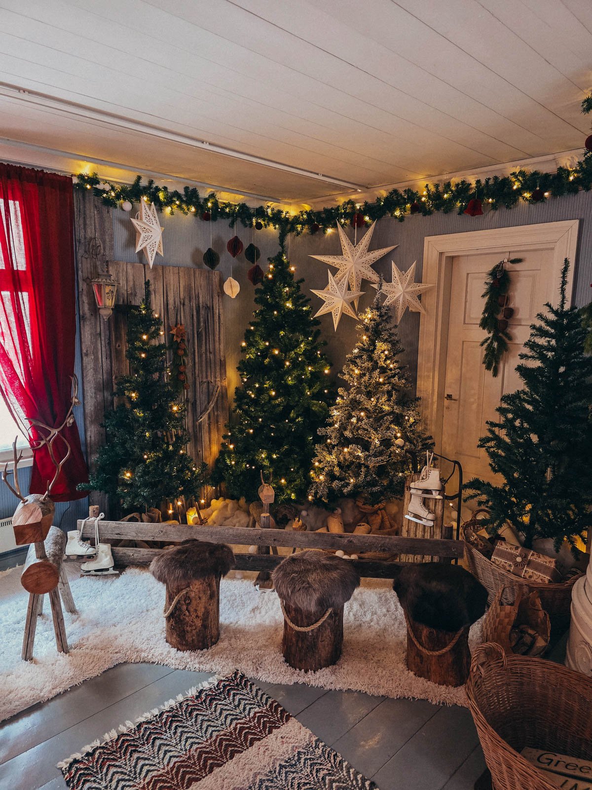 Three decorated Christmas trees inside a room with three wooden log seats covered in fur in front of the trees