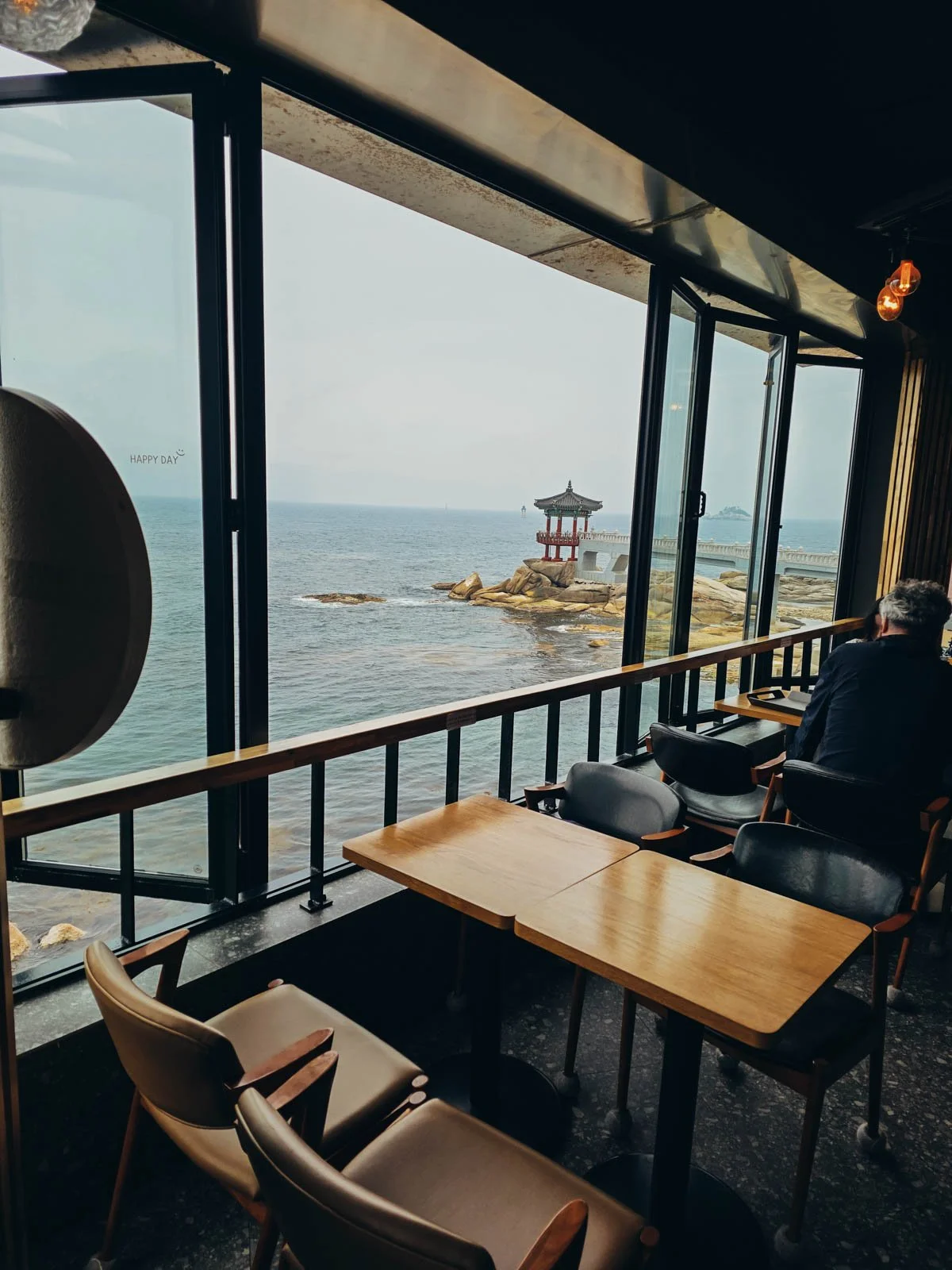 Tables at a waterfront cafe with windows open and overlooking the sea where a temple pavillion can be seen out on some rocks.