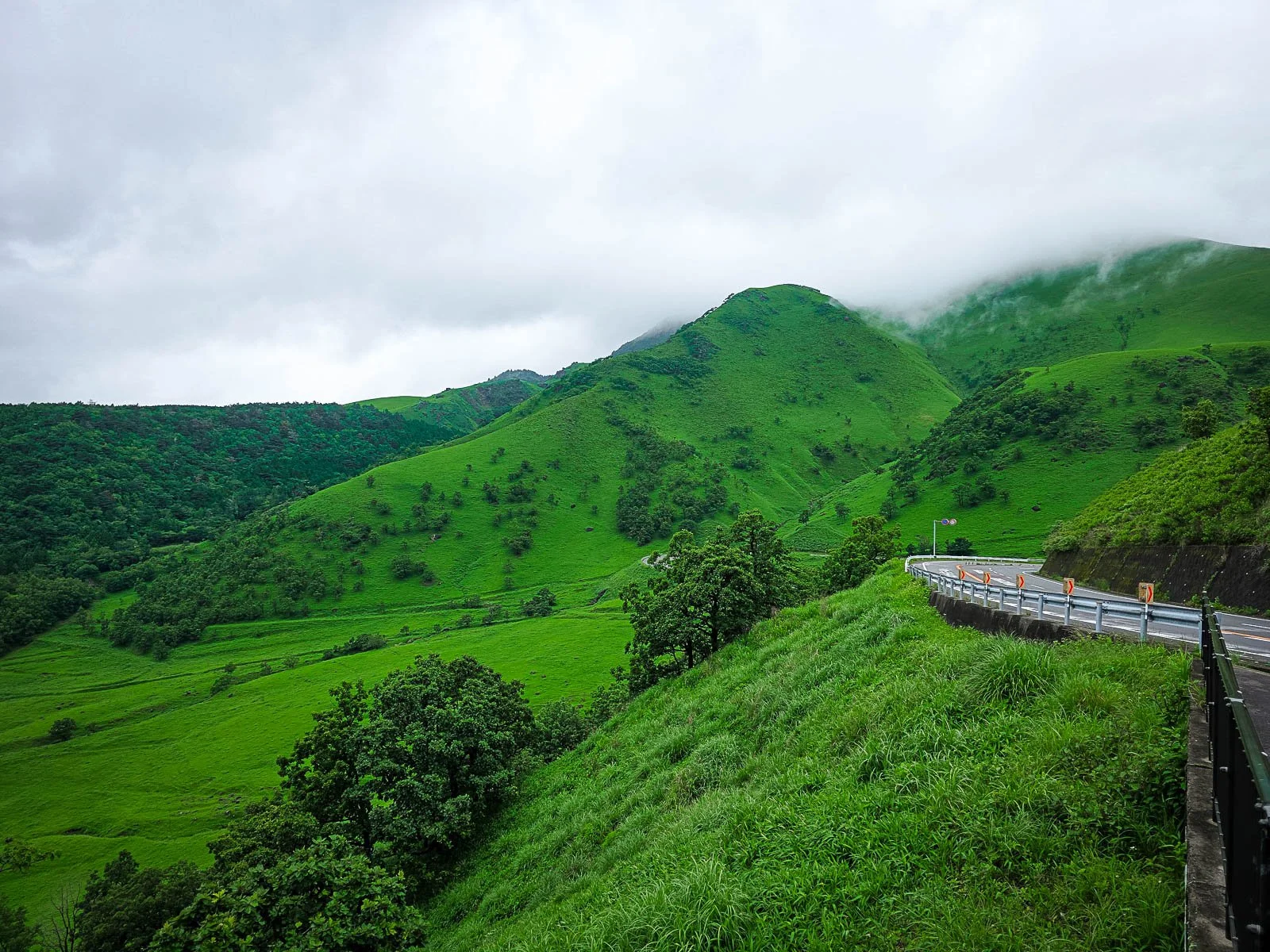 A lush green mountain landscape with low hanging clouds and a mountain road on the edge of the mountain to the right