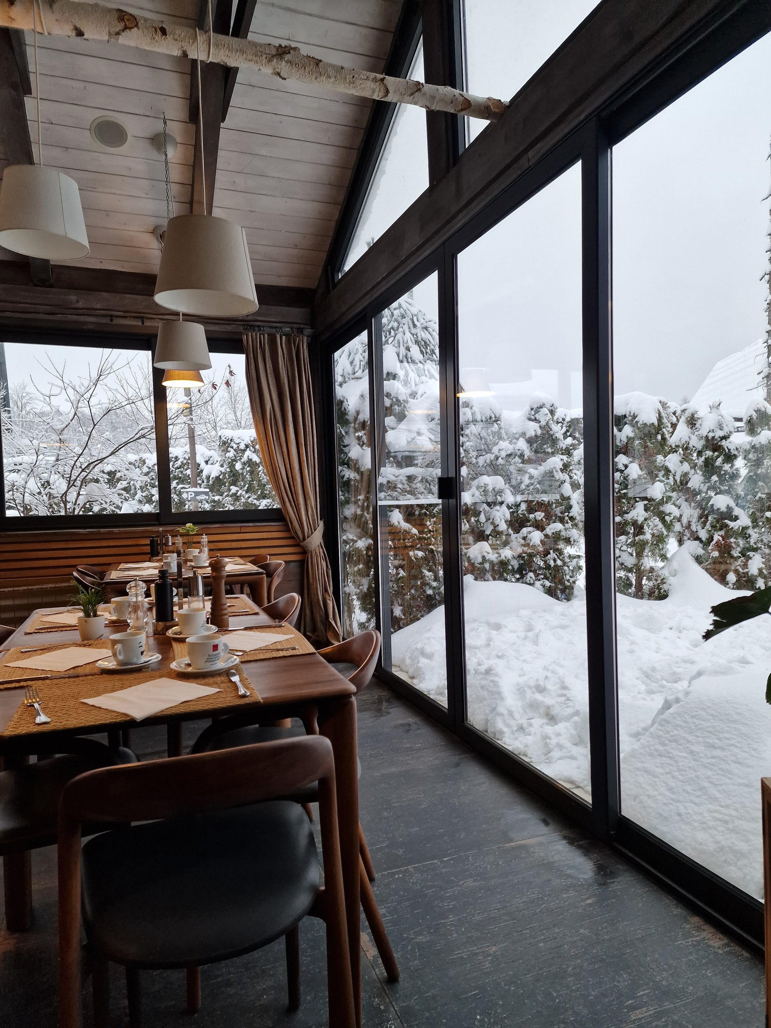A restaurant with a wooden table set for 6 people next to a huge floor to ceiling window where snow drifts can be seen outside