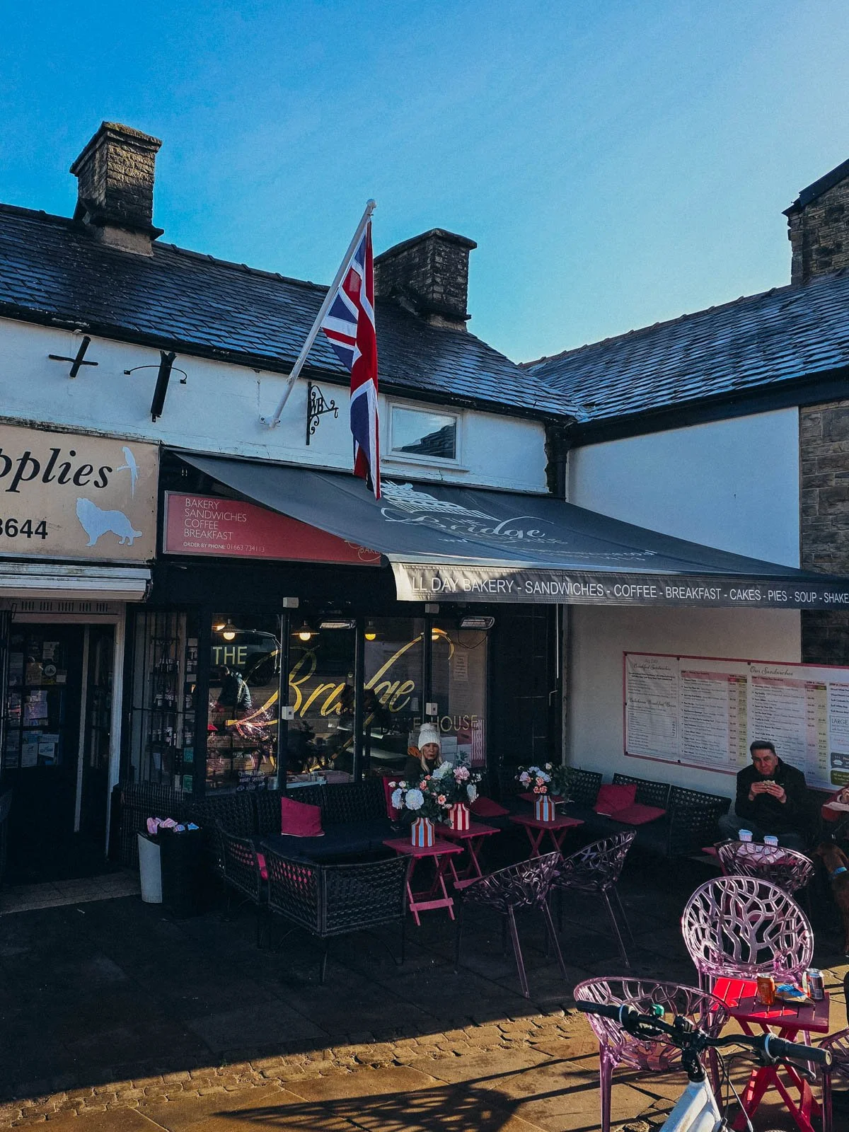 A small sandwich shop in the corner on the high street in Whaley Bridge with seating outside under an awning with a flag atached to the front