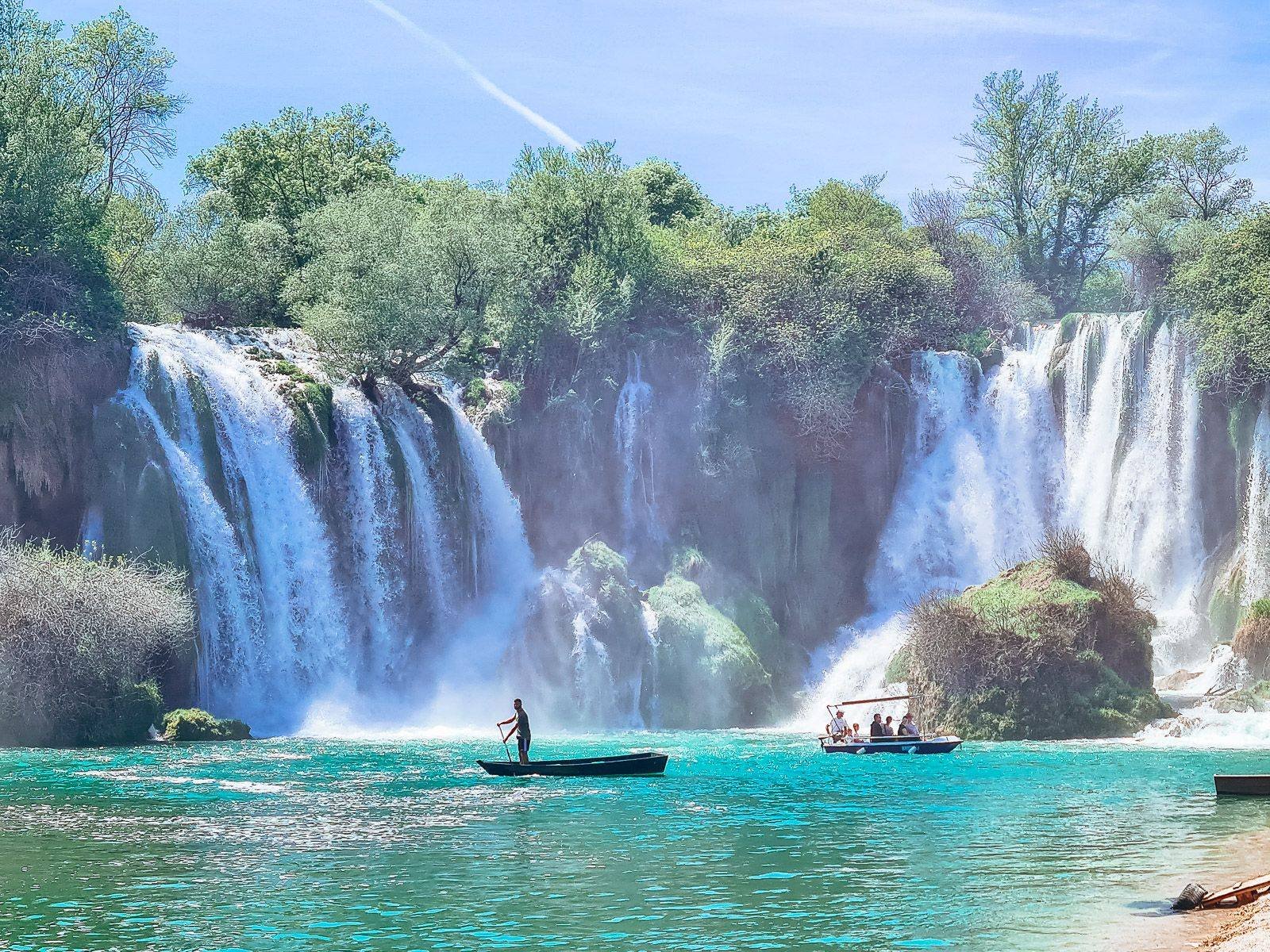 mist rising from a blue lake as waterfalls crash down around a man on a canoe