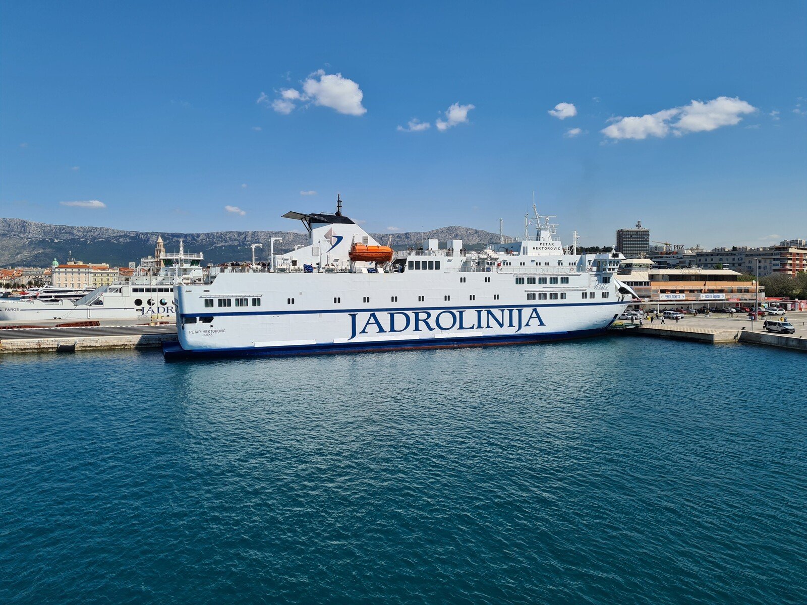 A large white ferry boat in the water docked at the port with blue writing that reads "Jadrolinija"