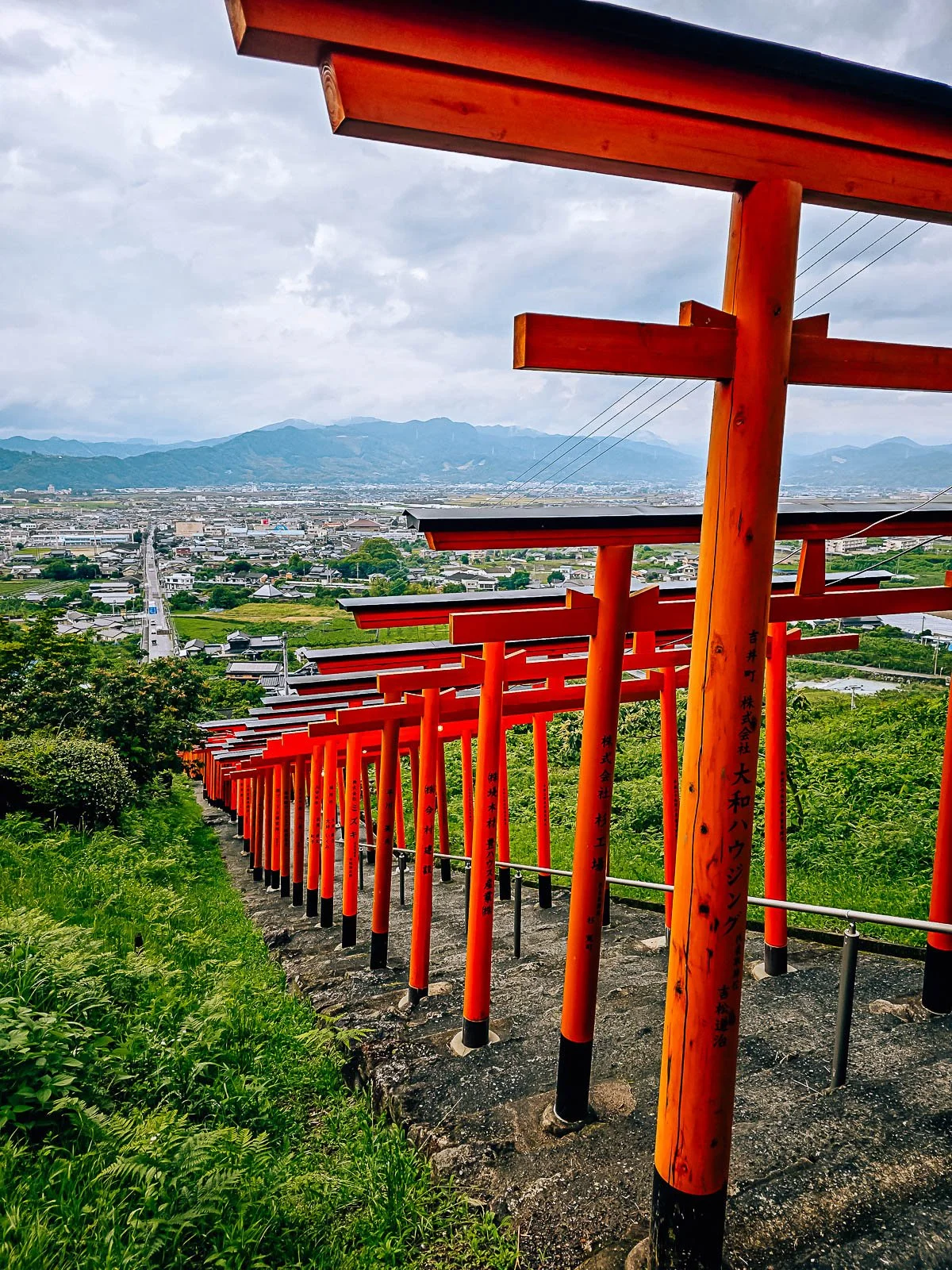 Many orang torii gates over stairs going down a green hill with a city in the far distance