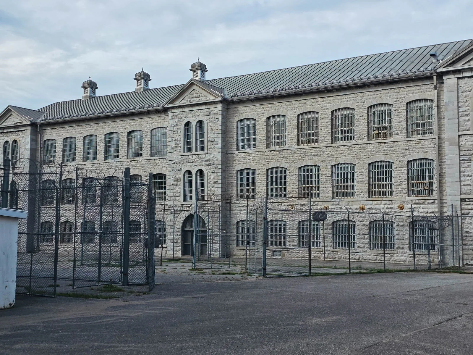 A large old stone penitentiary with many barred windows and a tall chain link fence in front of the building
