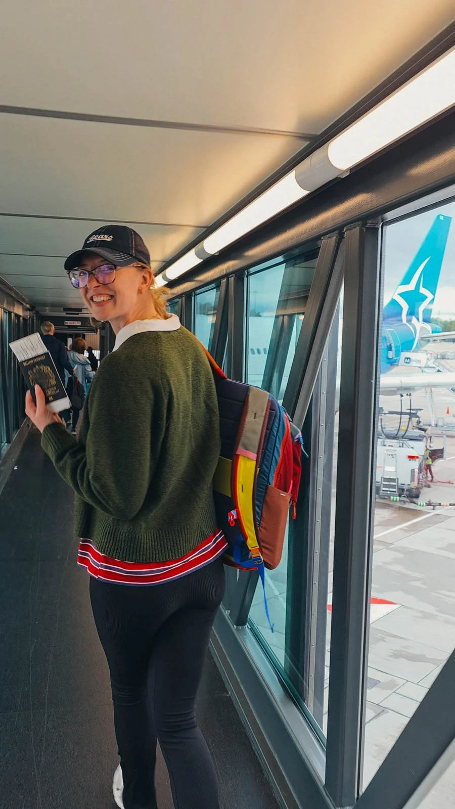 Helena walking down a jet bridge looking back over her shoulder smiling and holding her passport up to the camera. She is wearing black trousers and a green jumper with a colourful backpack over one shoulder. Out the window is an Air Transat airplane