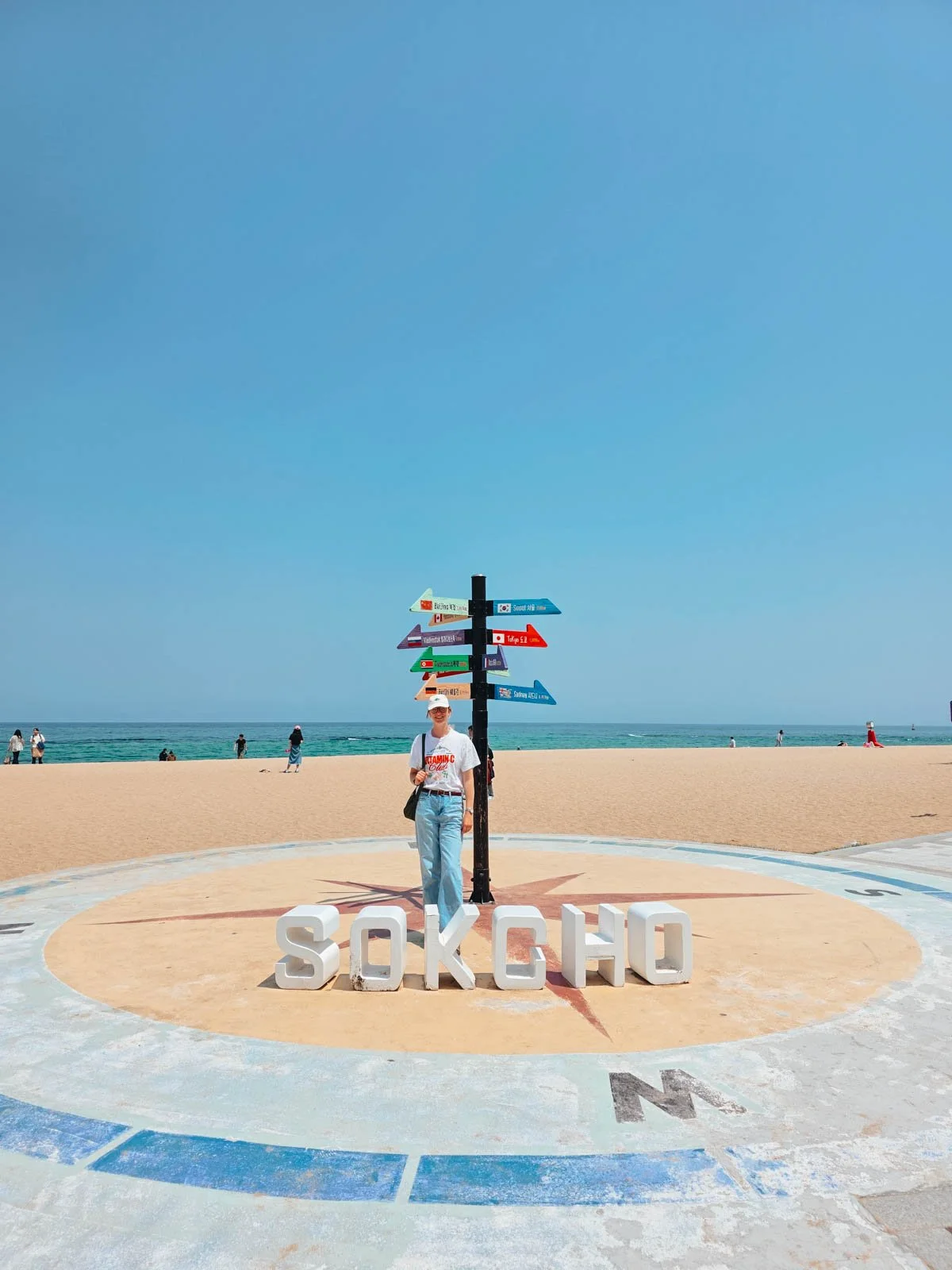 Helena in jeans and a white tshirt standing behind a white sign that says "Sokcho" and next to a signpost with colourful arrows pointing in lots of different directions. Behind is a sandy beach and it's a sunny day