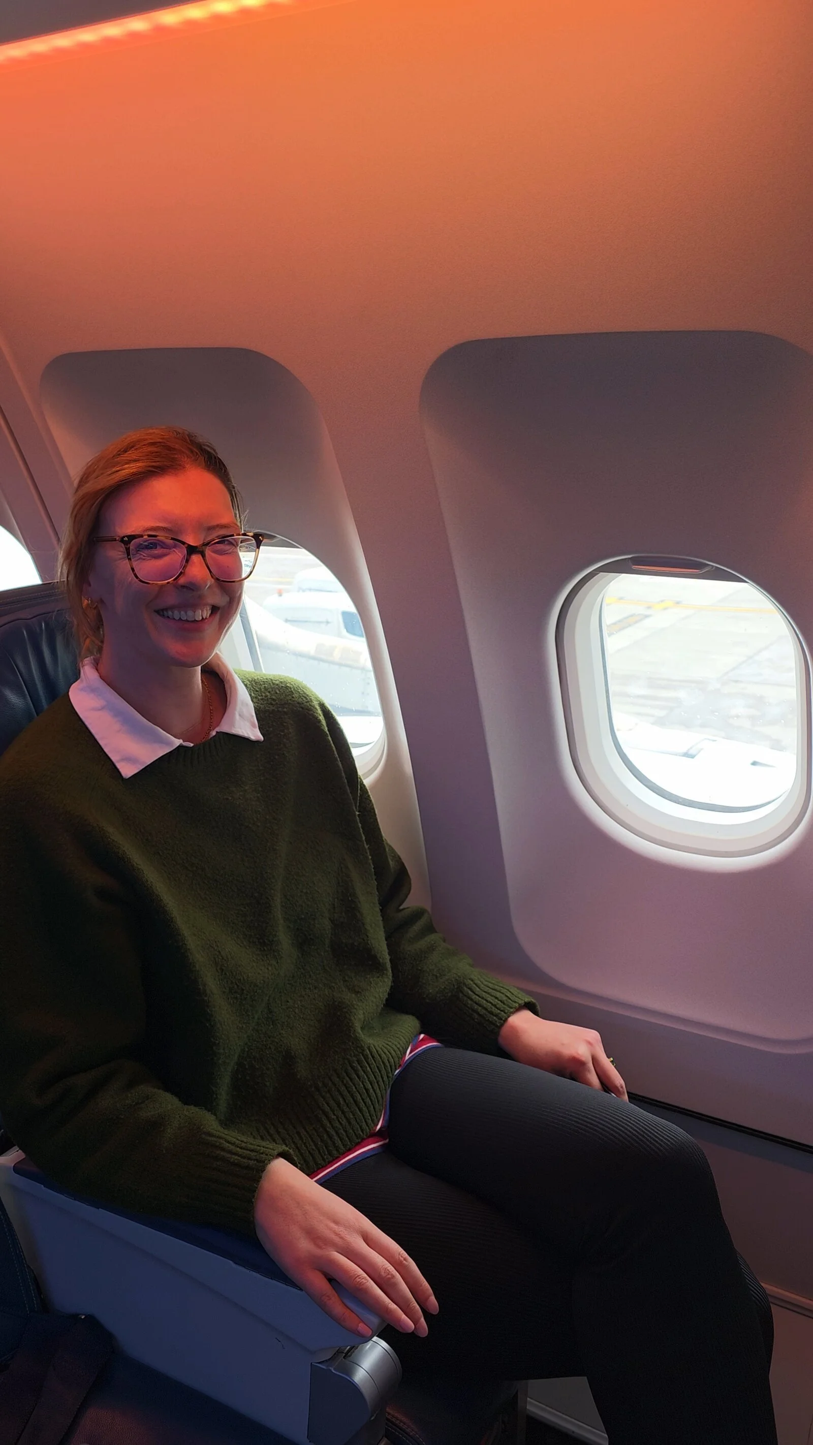 Helena in a green jumper and blac trousers sitting in a plane seat next to the window and smiling at the camera