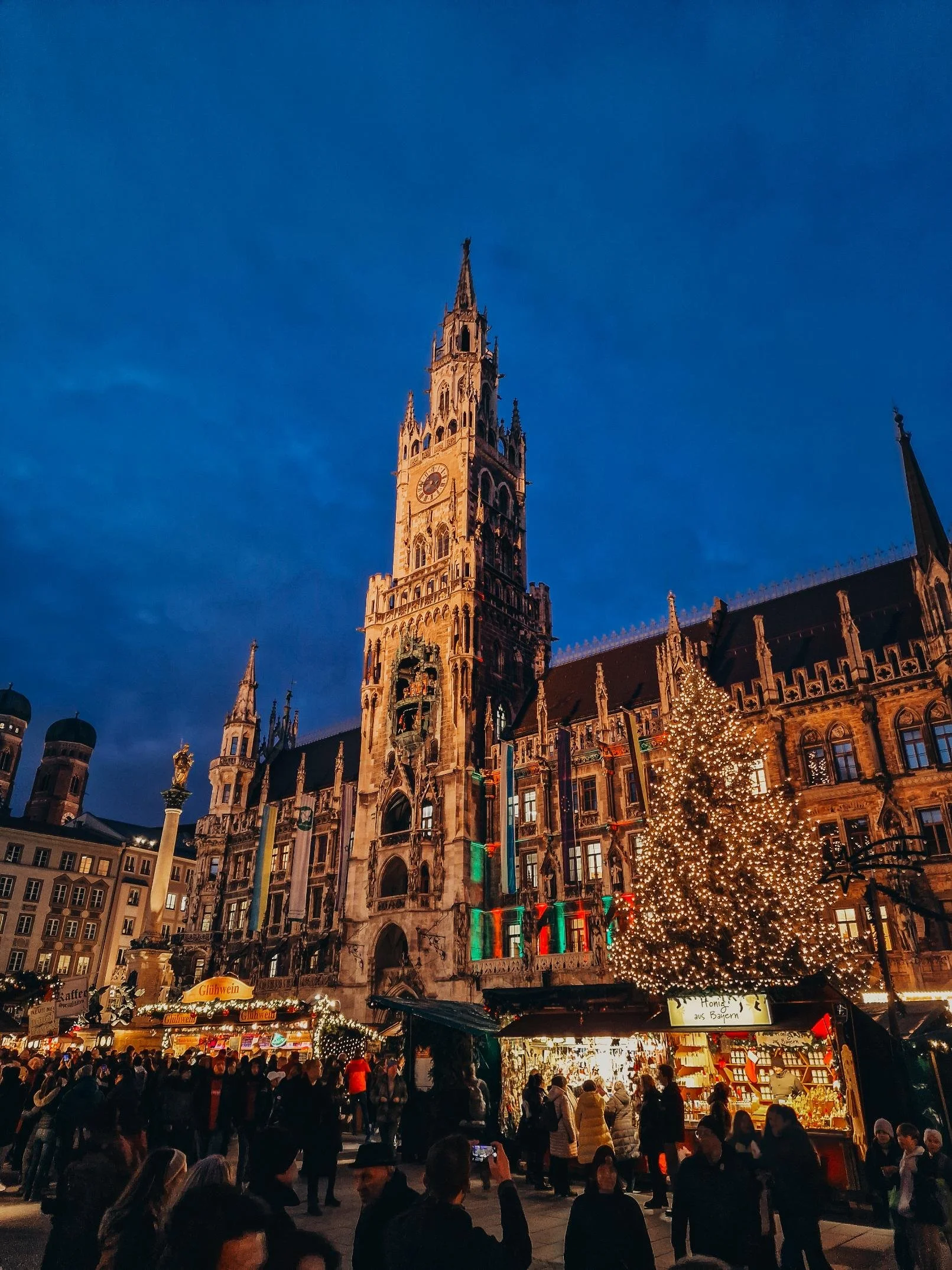 A large old ornate towered building decorated for christmas with a busy market area, The market area has may little stalls a a large lit Christmas Tree at night
