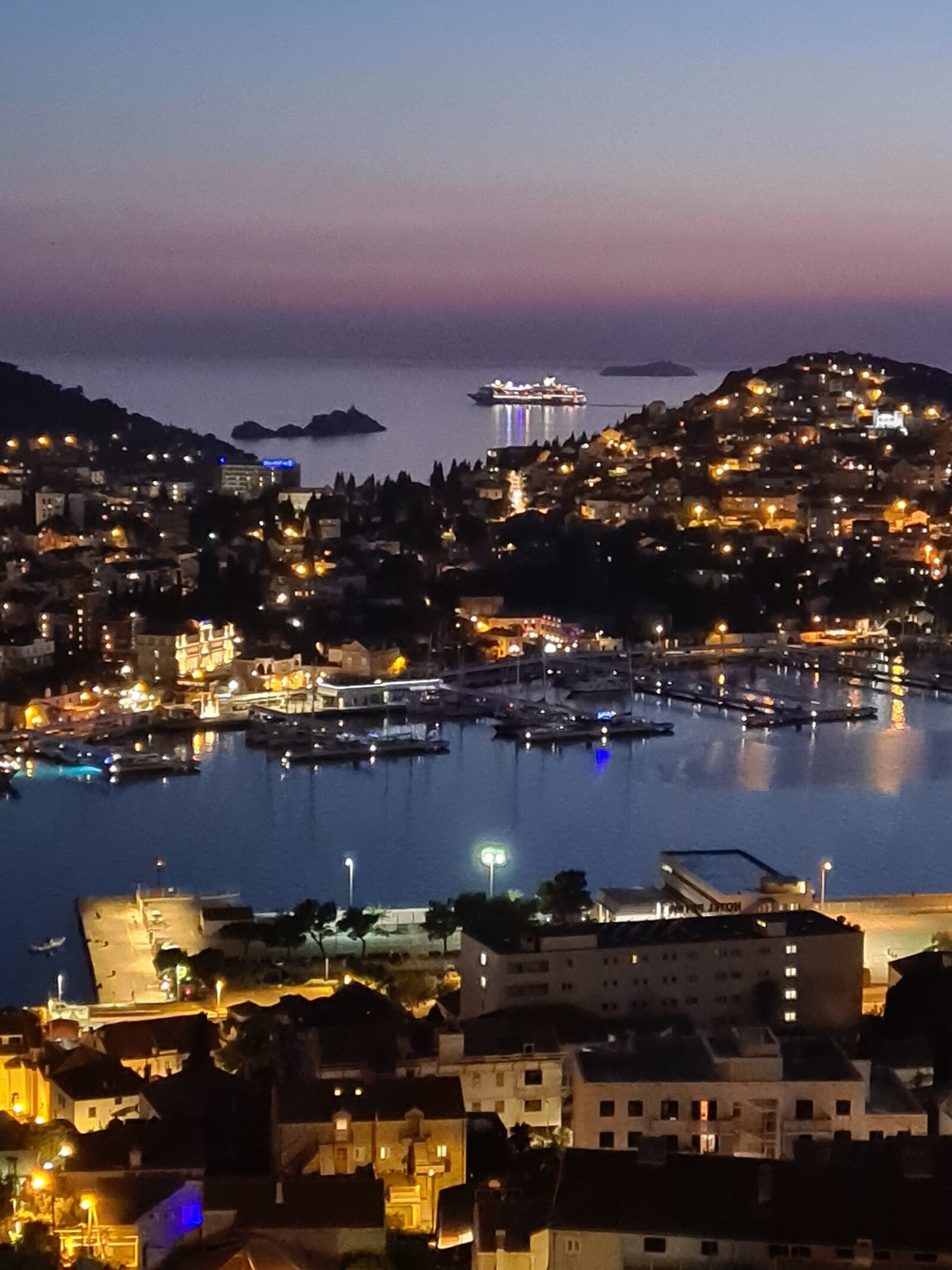 A view of hills between two bodies of water with many lit up houses and a marina at night. A lit up cruise ship is in the distance
