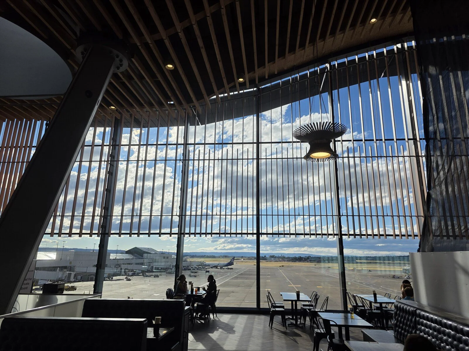 Inside a large airy bar at at PDX airport with floor to ceiling windows offering a view of planes on the tarmac. Two people are sitting at tables silhouetted by the window