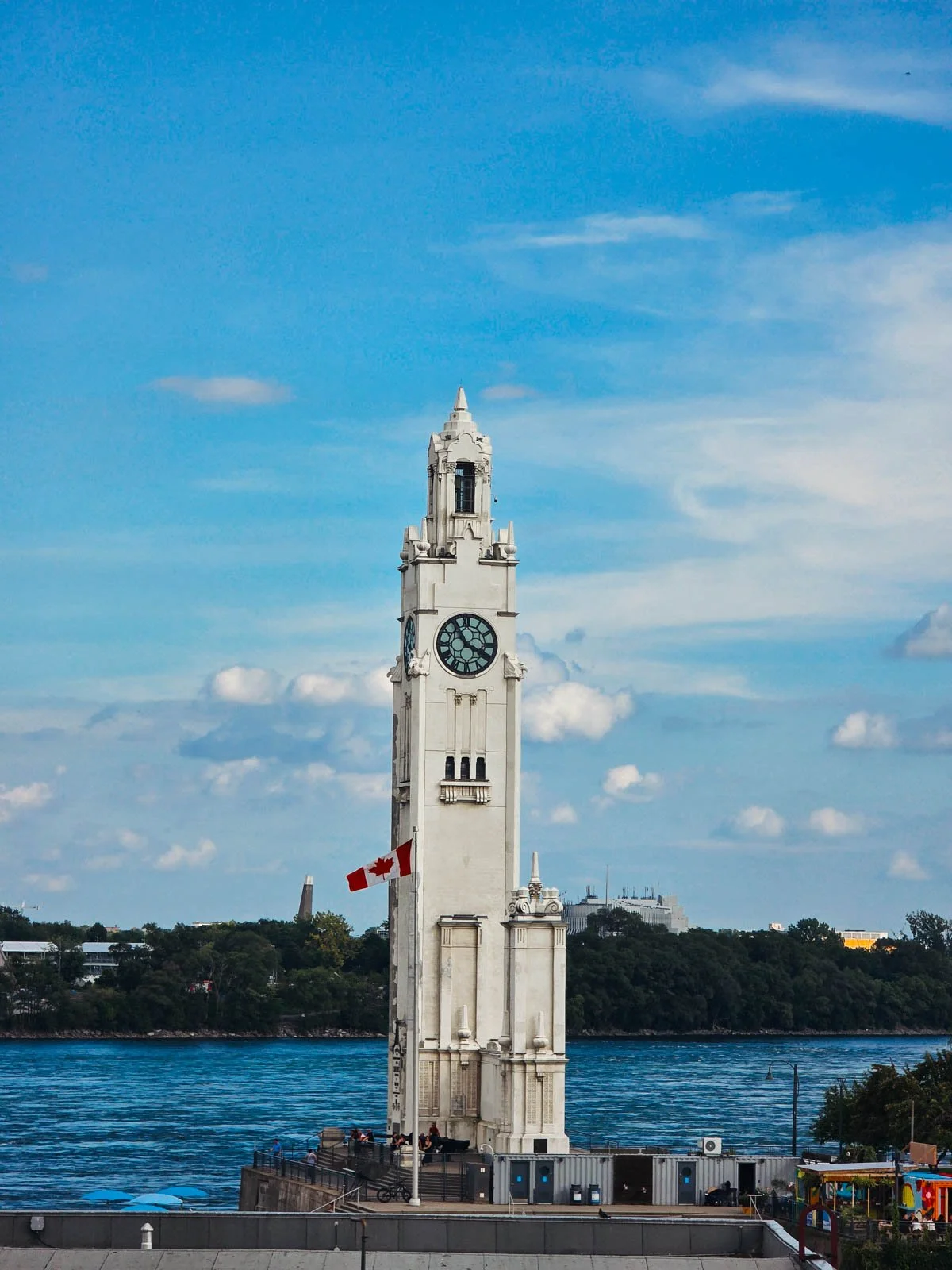 A white clock tower along the waterfront with the Canadian flag flying in front of it