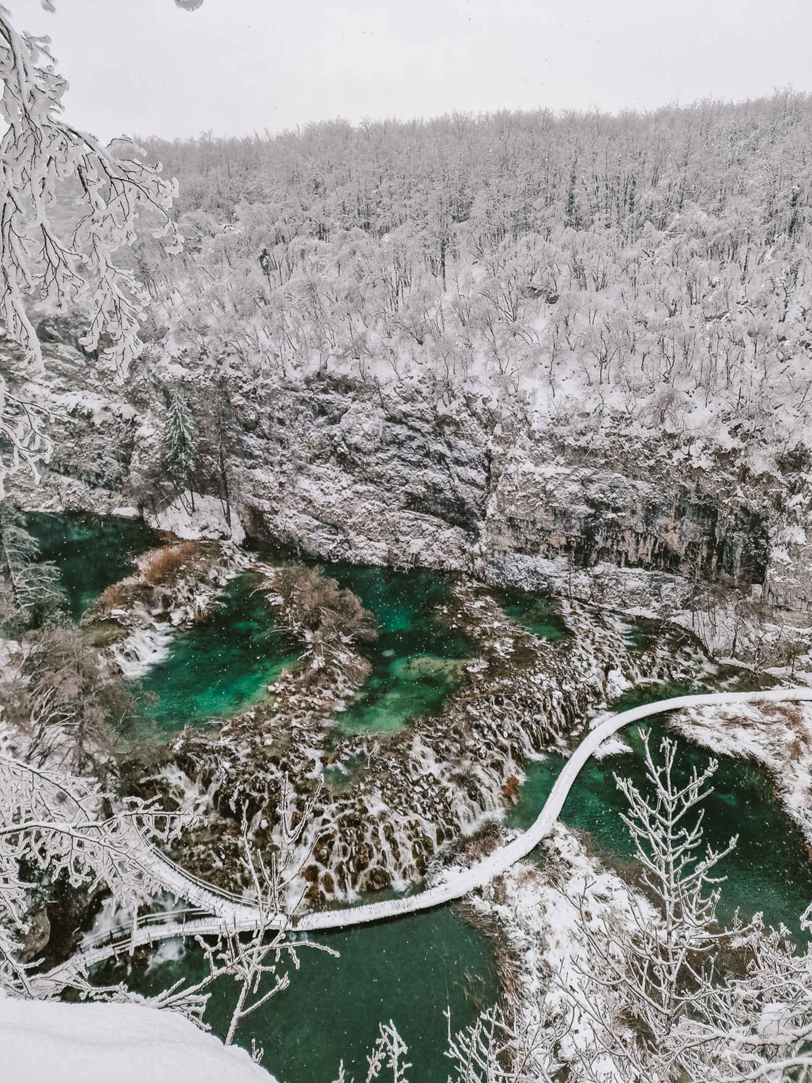 View from above on a canyon trail looing down into the canyon where green lakes flow over rocks and around a snow covered walkway with snowy trees and cliffs either side of the water in Plitvice Lakes National Park, Croatia in winter