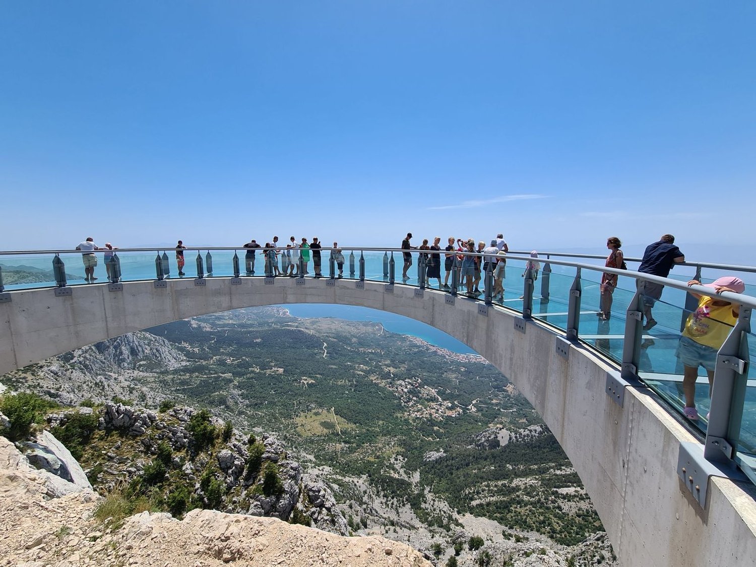 a glass bridge arching out from the mountain edge with people standing on it and adrop below with a view of the coast in the distance