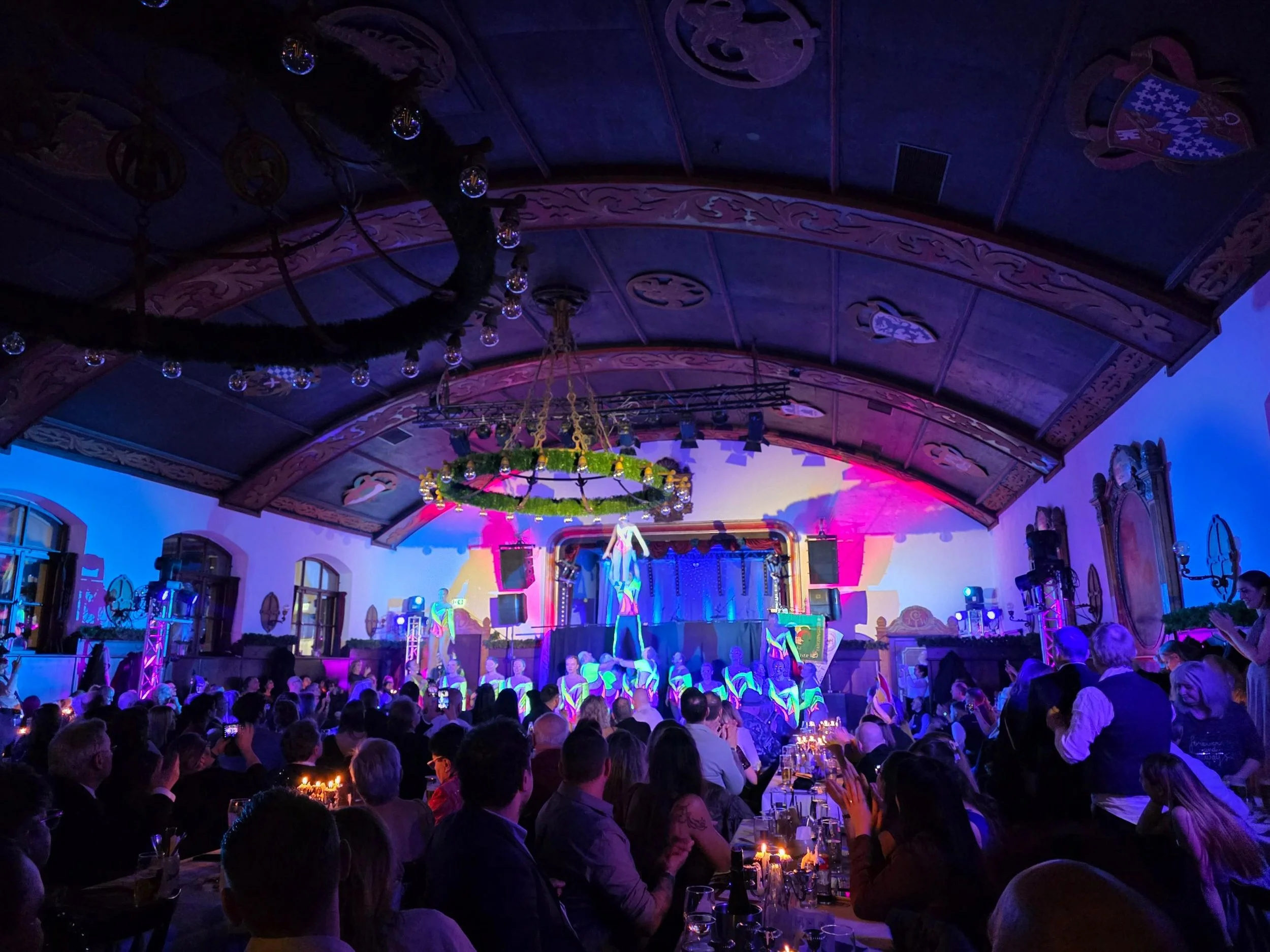 many people sitting along long tables watching a performance in a dark ornate beer hall