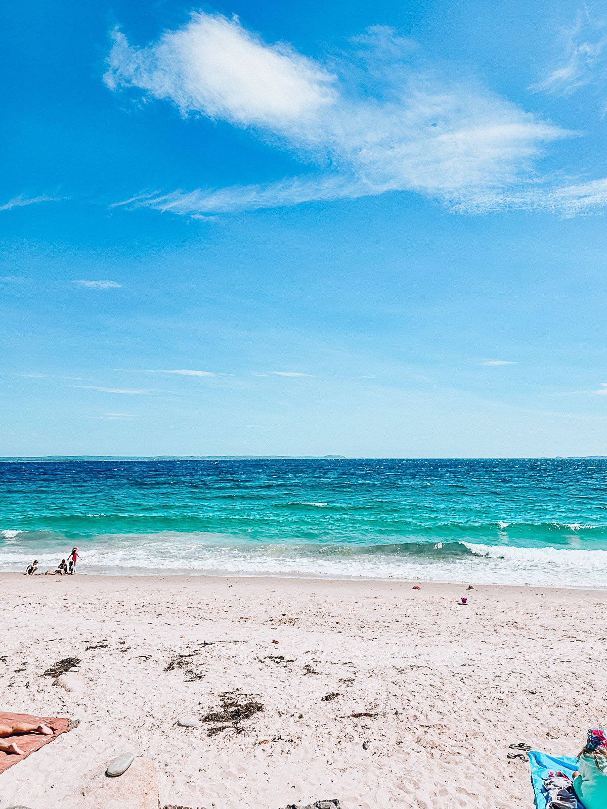 A white sandy beach on a clear sunny day with small turquoise waves crashing along the shore where a group of children play in the distance