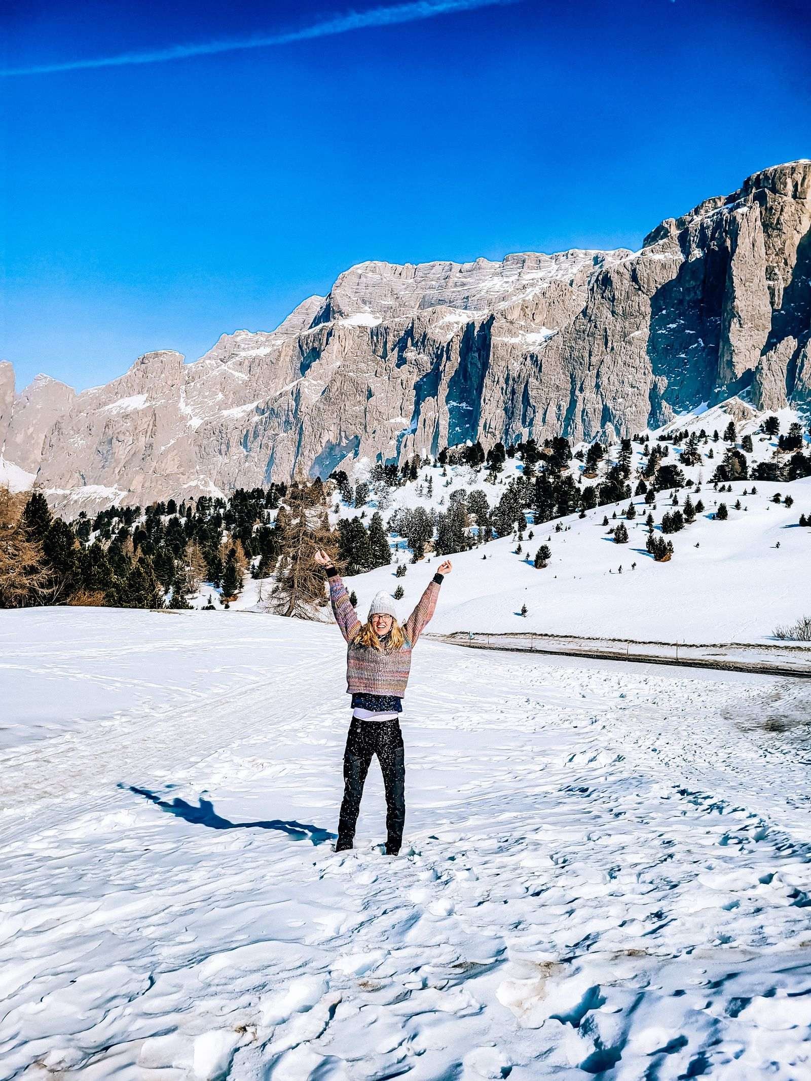 A girl standing in snow with her hands up in the air. Many snow coved jagged Dolomite mountains are in the background
