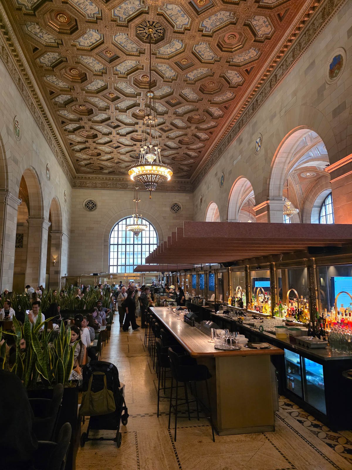Inside an ornate room with huge high ceilings and an ornate ceiling decor with massive chandeliers and stone arches along each side of the room with huge domed window at the end. Once a bank and now a coffee shop with many people sitting at tables