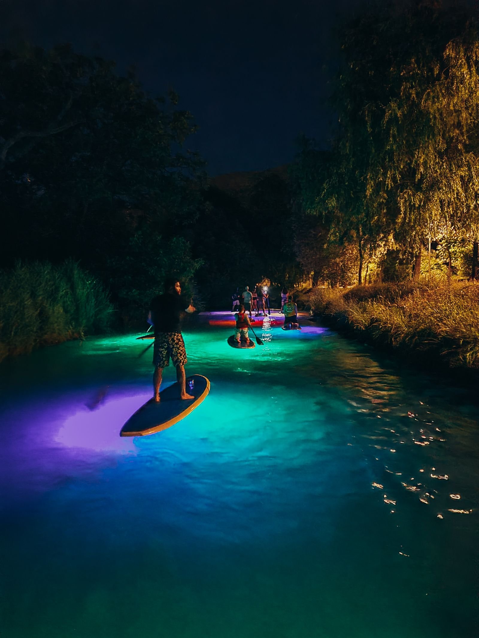 Many people standing on paddleboard lit up in neon colours going down a river at night