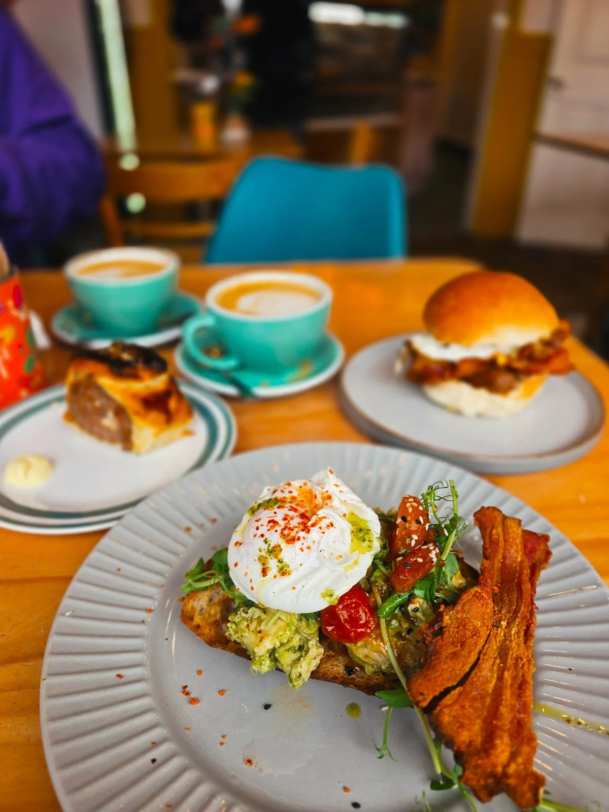 Three plates of food on a table, the closest is avocado on toast with bacon and poached egg, behind is a breakfast sandwich, a sausage rolls and two cups of coffee in turquoise cups - the perfect start to your High Peak day trip from Manchester