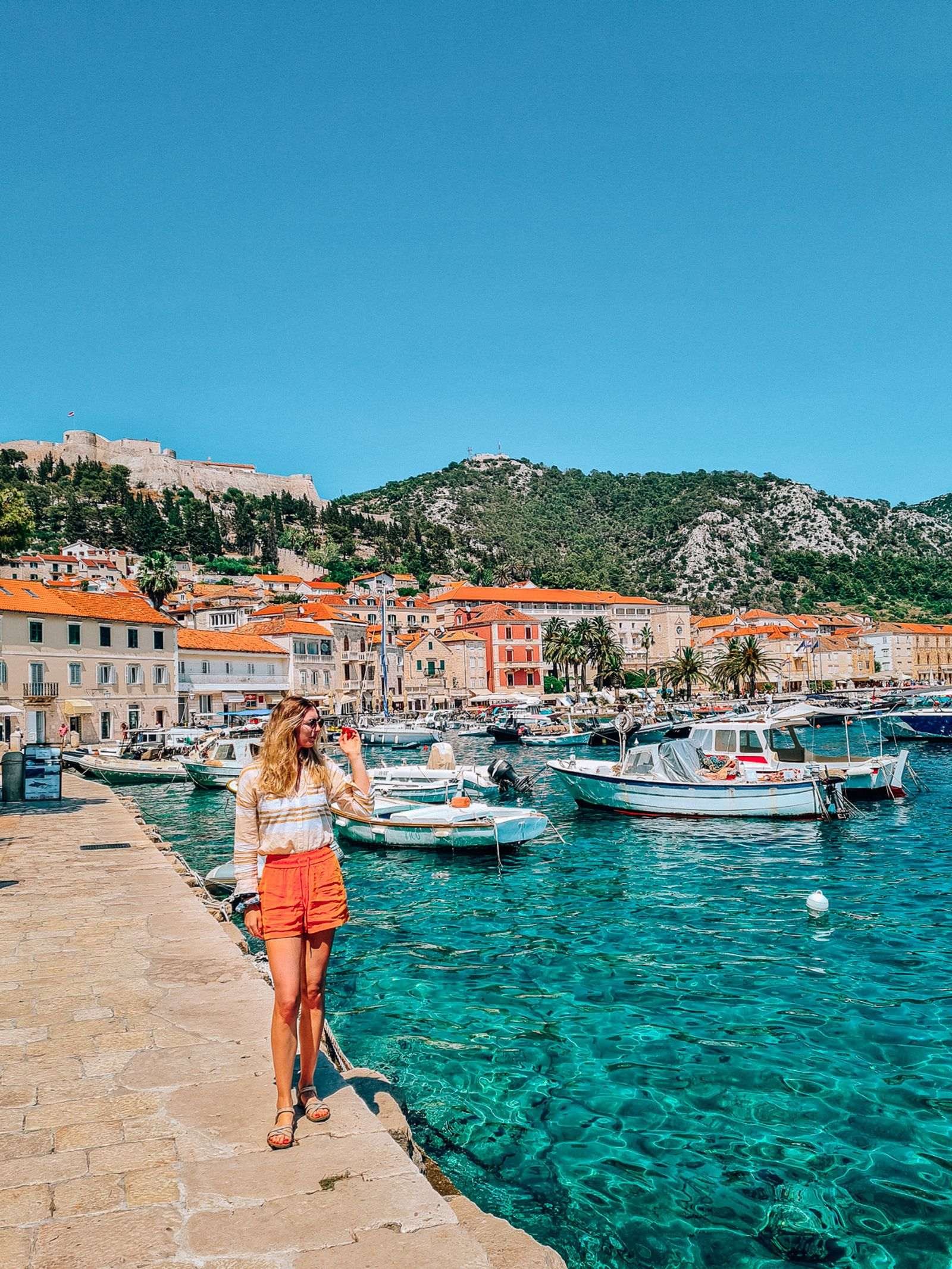 girl in orange shorts and yellow shirt standing on a harbour wall with view of hvar old town stone buildings and a fortress on the hill in the background