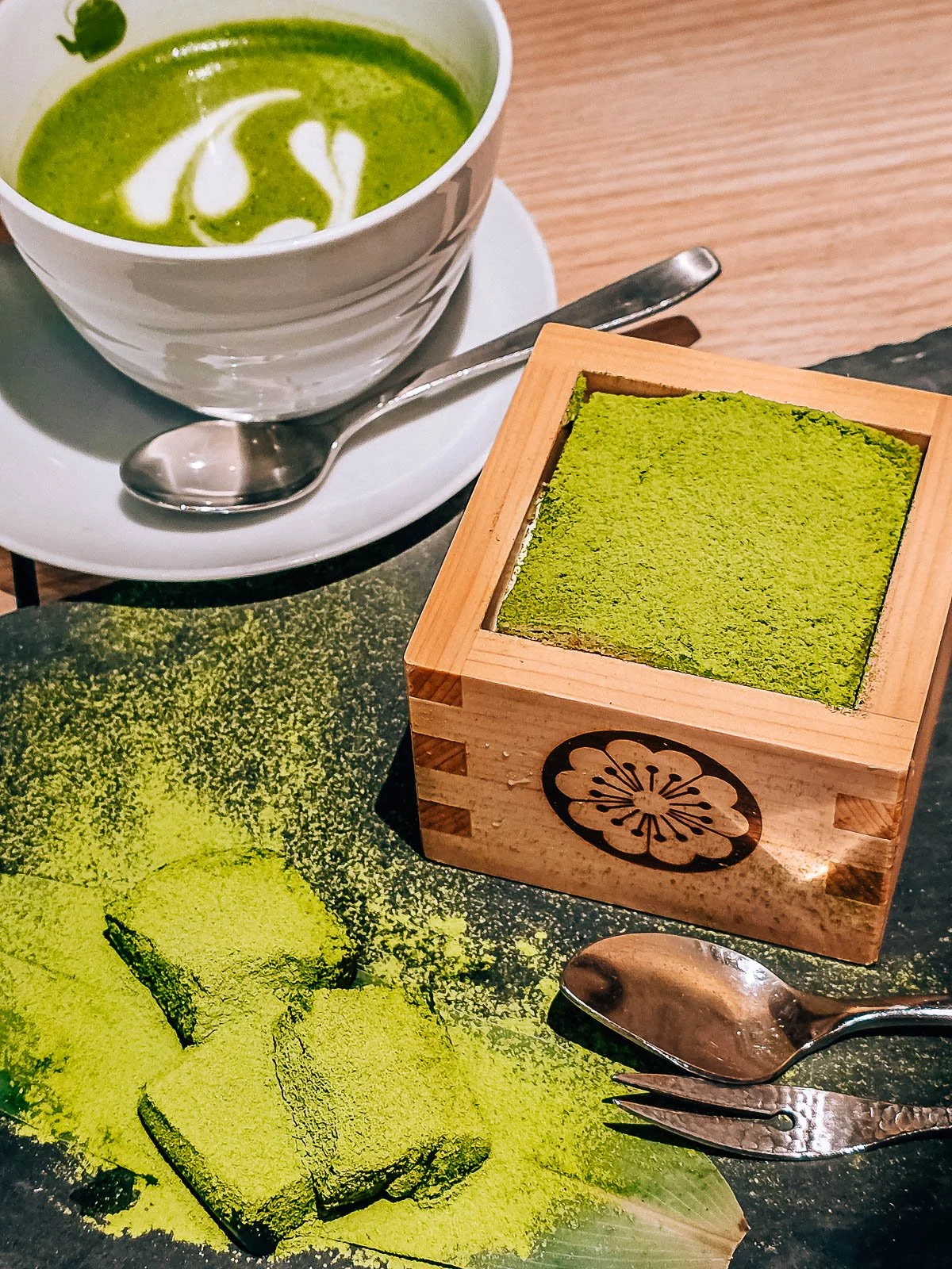 A vibrant green matcha coffee in a white cup next to a green match desert in square wooden bowl on a table