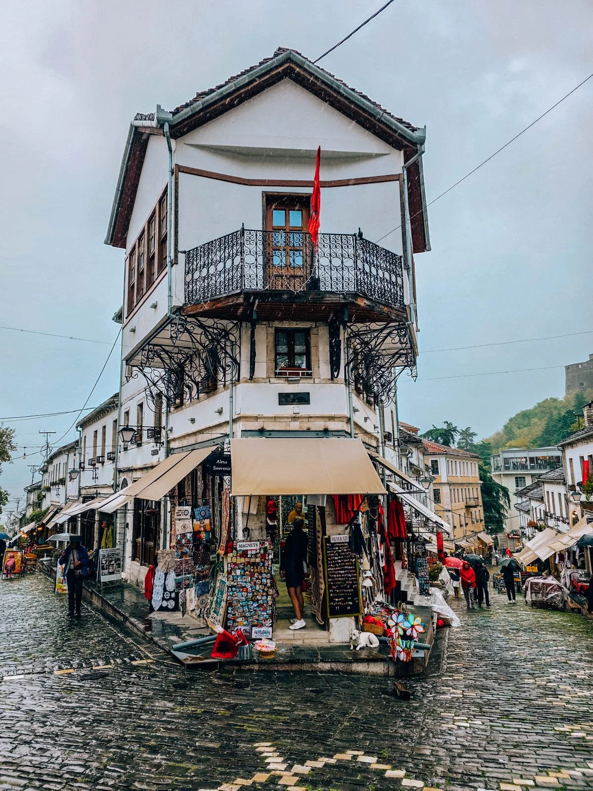 A white shop at the corner of a cobbled street in an old town. Many shops line each side of the street with loads of trinkets and sale items lining the street