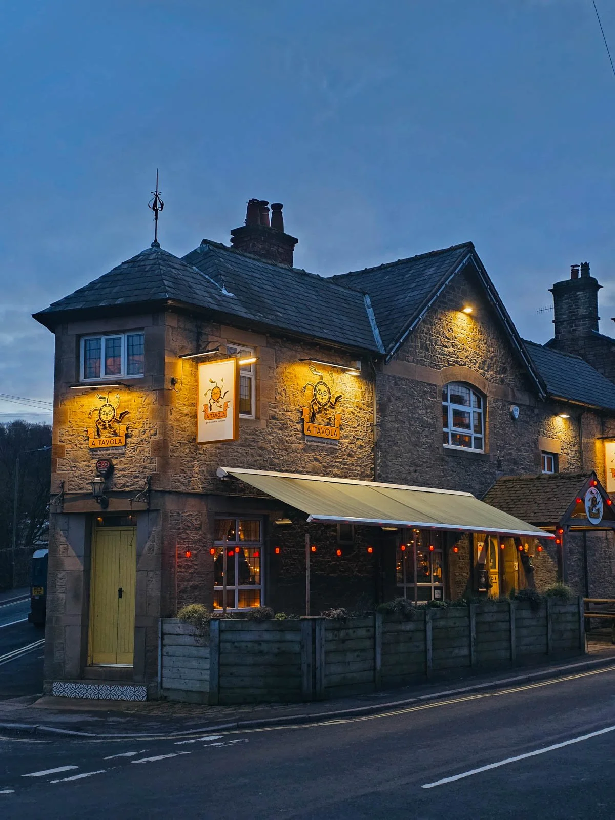 a cosy looking stone building which houses a Sicilian restaurant in New Mills, High Peak. The restaurant signs are lit by warm yellow light and the front door is yellow