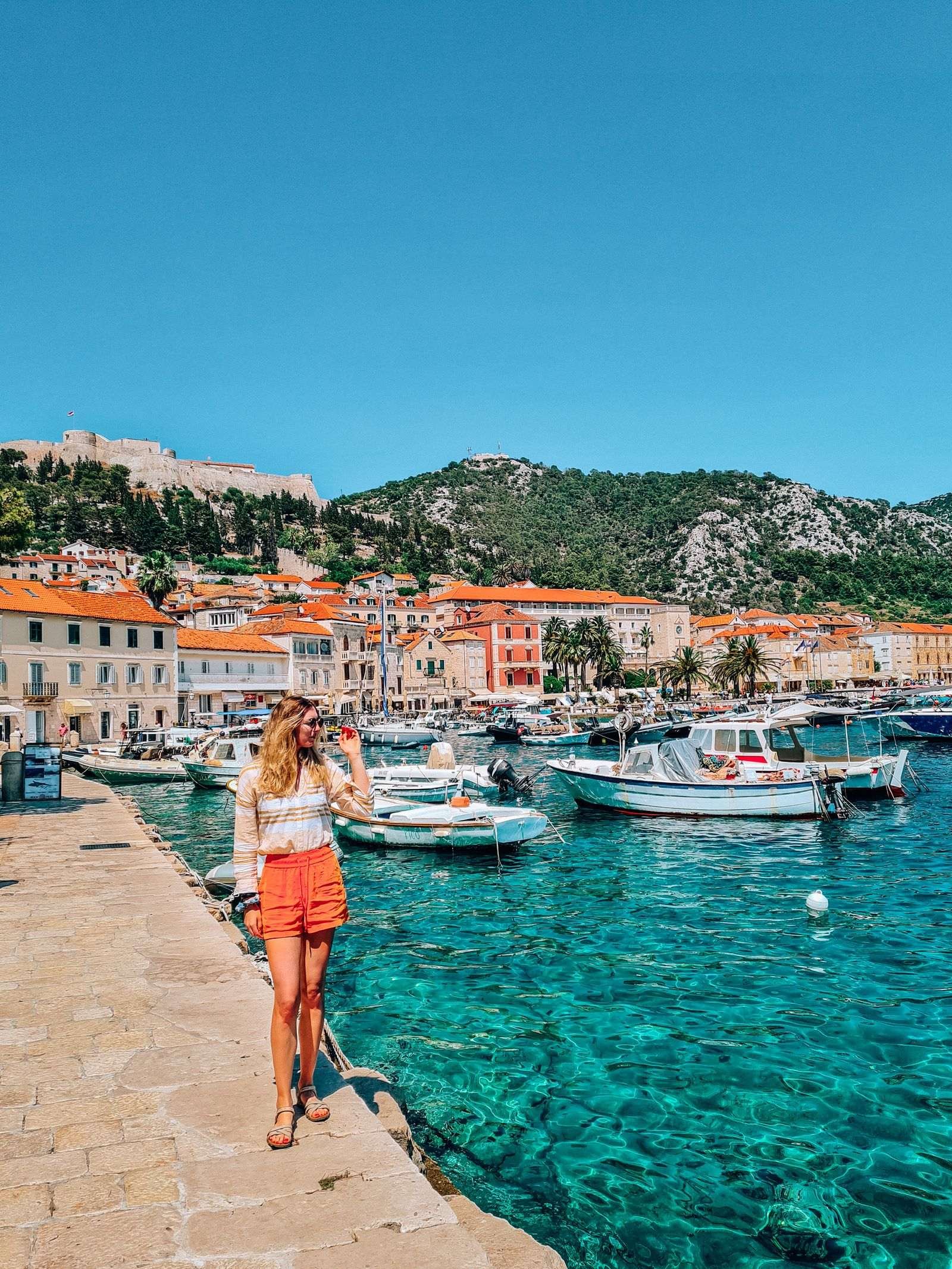 A girl in orange shorts standing on the edge of the clear blue harbour of Hvar with many white boats and stone buildings in the background