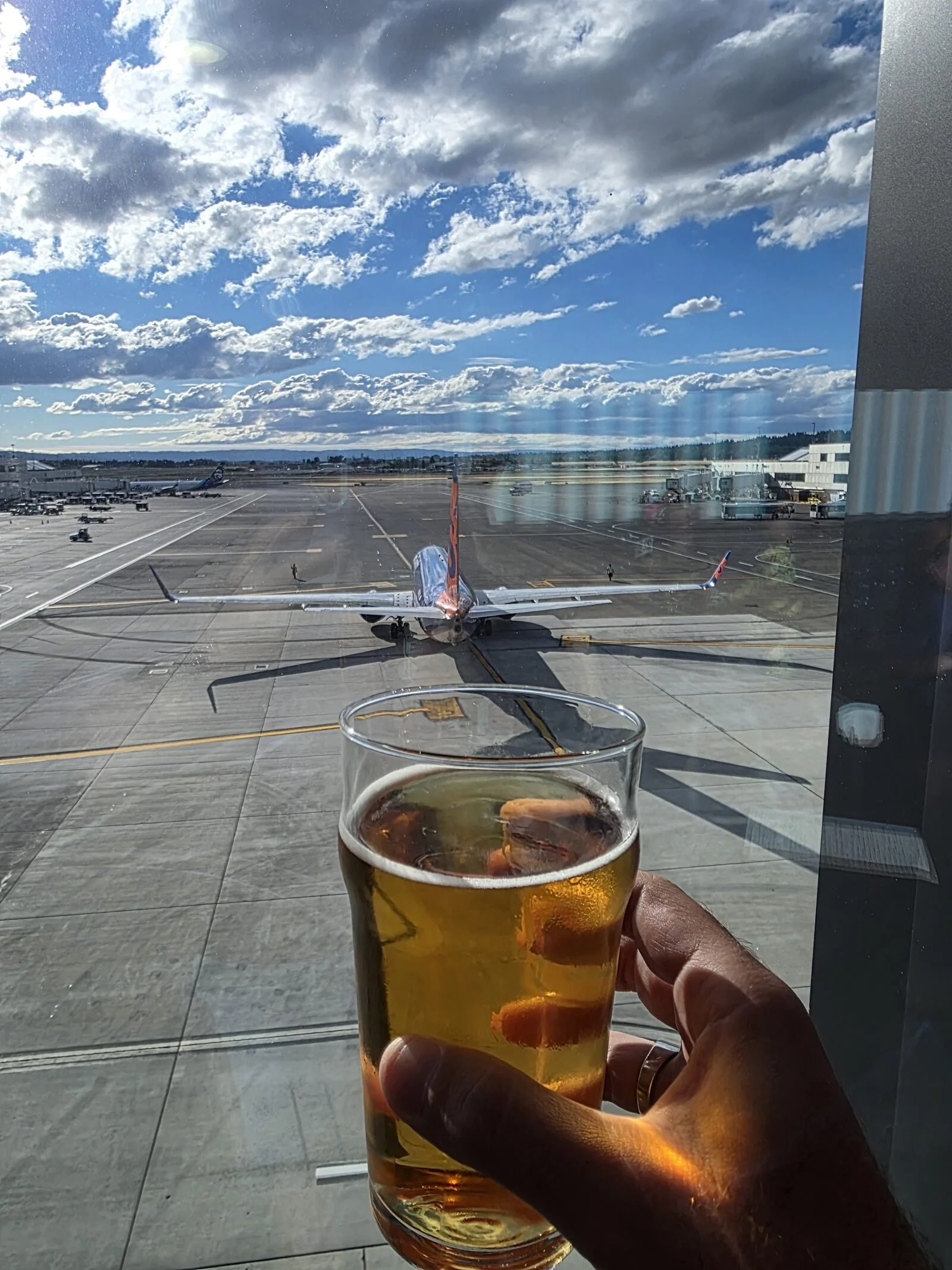 Holding a pint of beer up to a large window where there is a view of a plane taxiing on the tarmac at an airport