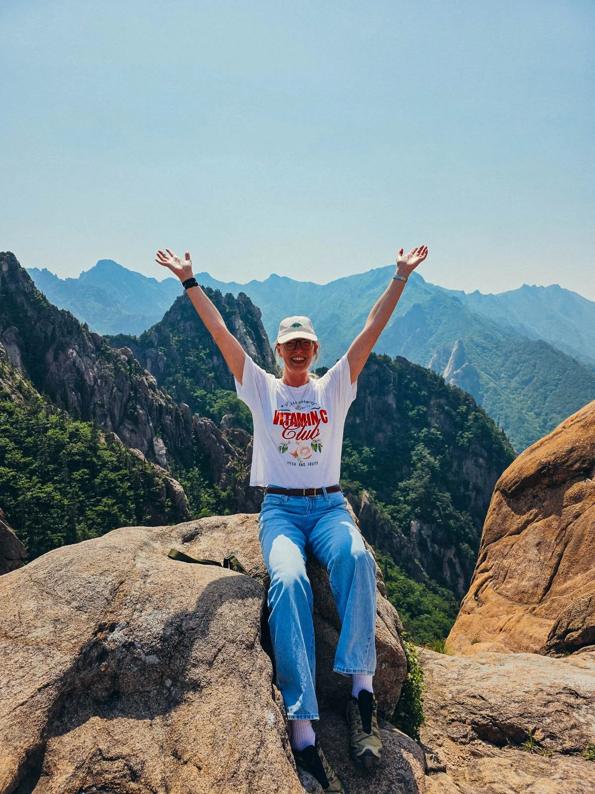 Helena in jeans and a white tshirt, sitting on a rock with her arms in the air and a rocky, vast landscape behind her