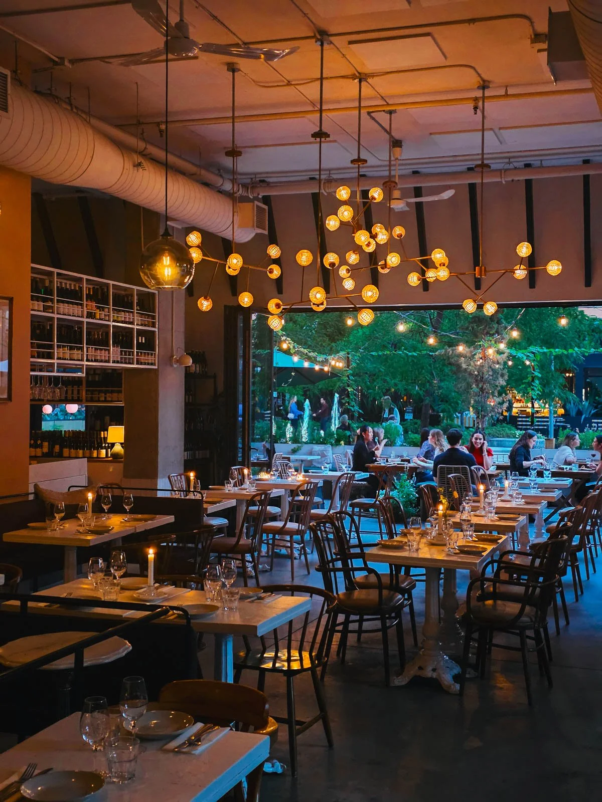 The interior of a restaurant with many tables set and warm light bulbs hanging from the ceiling, at the far end of the room the whole wall is open to a terrace where people are eating