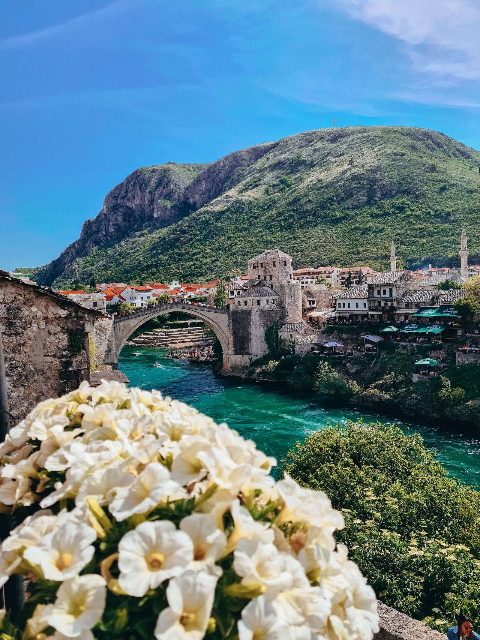 A closeup picture of white flowers with a stone bridge crossing over a blue river and many stone buildings in the background next to a large hill