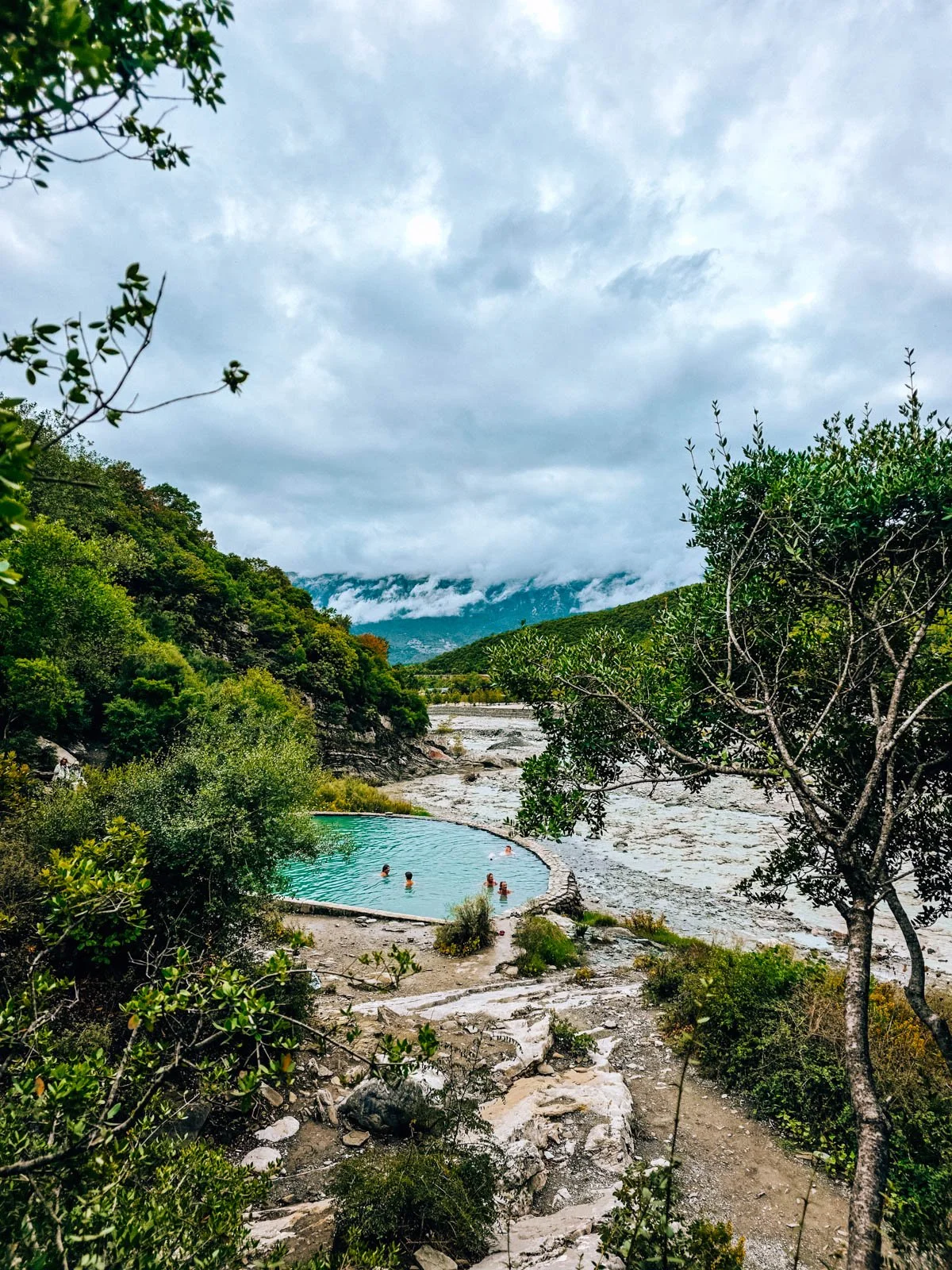 Bright blue thermal pools made from stone with people relaxing inside them outdoors surrounded by green trees