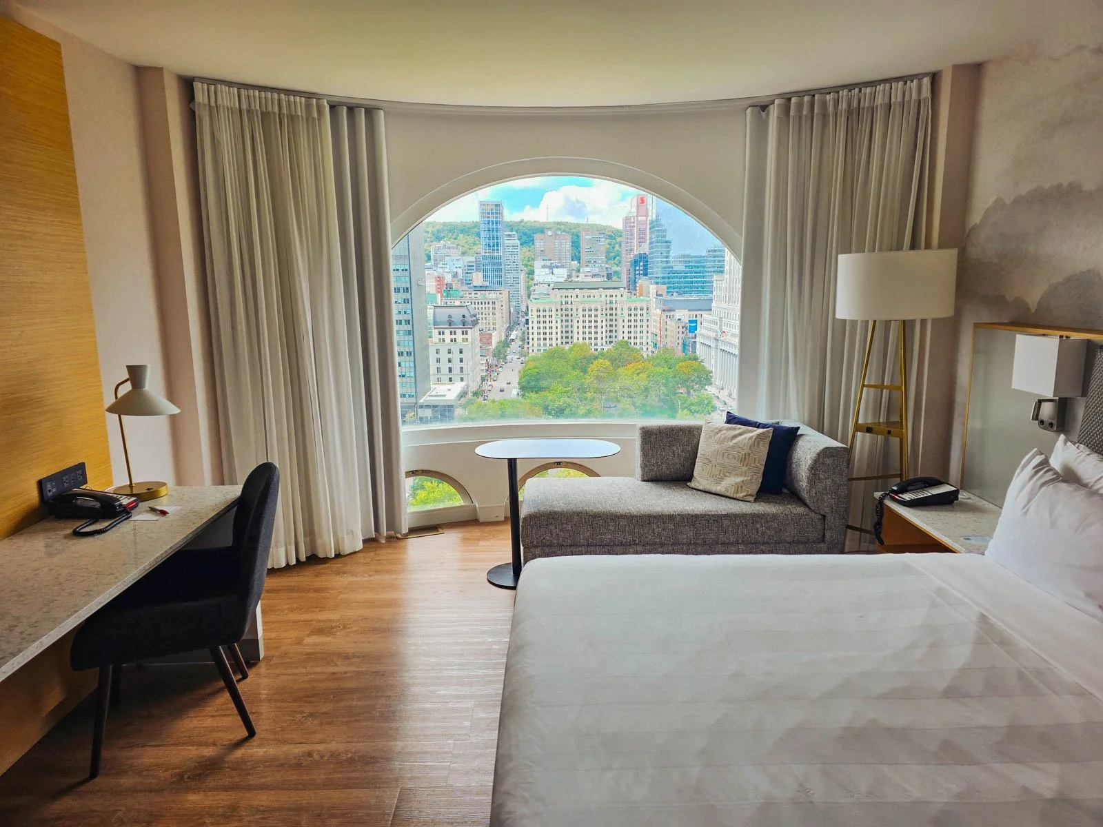 Out hotel room in Montreal with a large bed in the foreground, a desk against the left wall and a chais lounge in front of the domed window which has a view of downtown Montreal