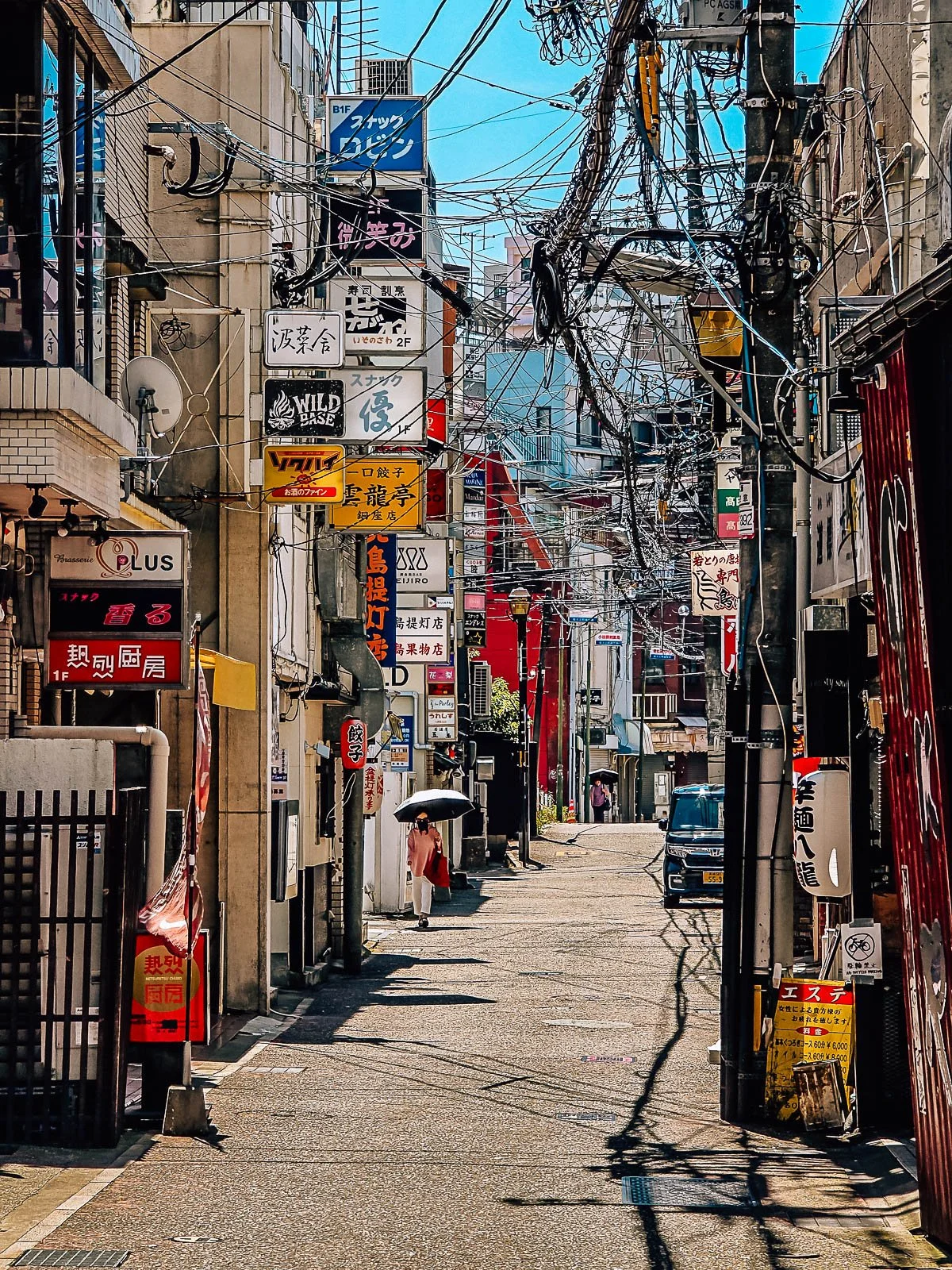 A narrow street lined with many colourful shop signs and lights. The street has hundreds of electrical wires drapped across it connected to an power pole in the foreground of the photo