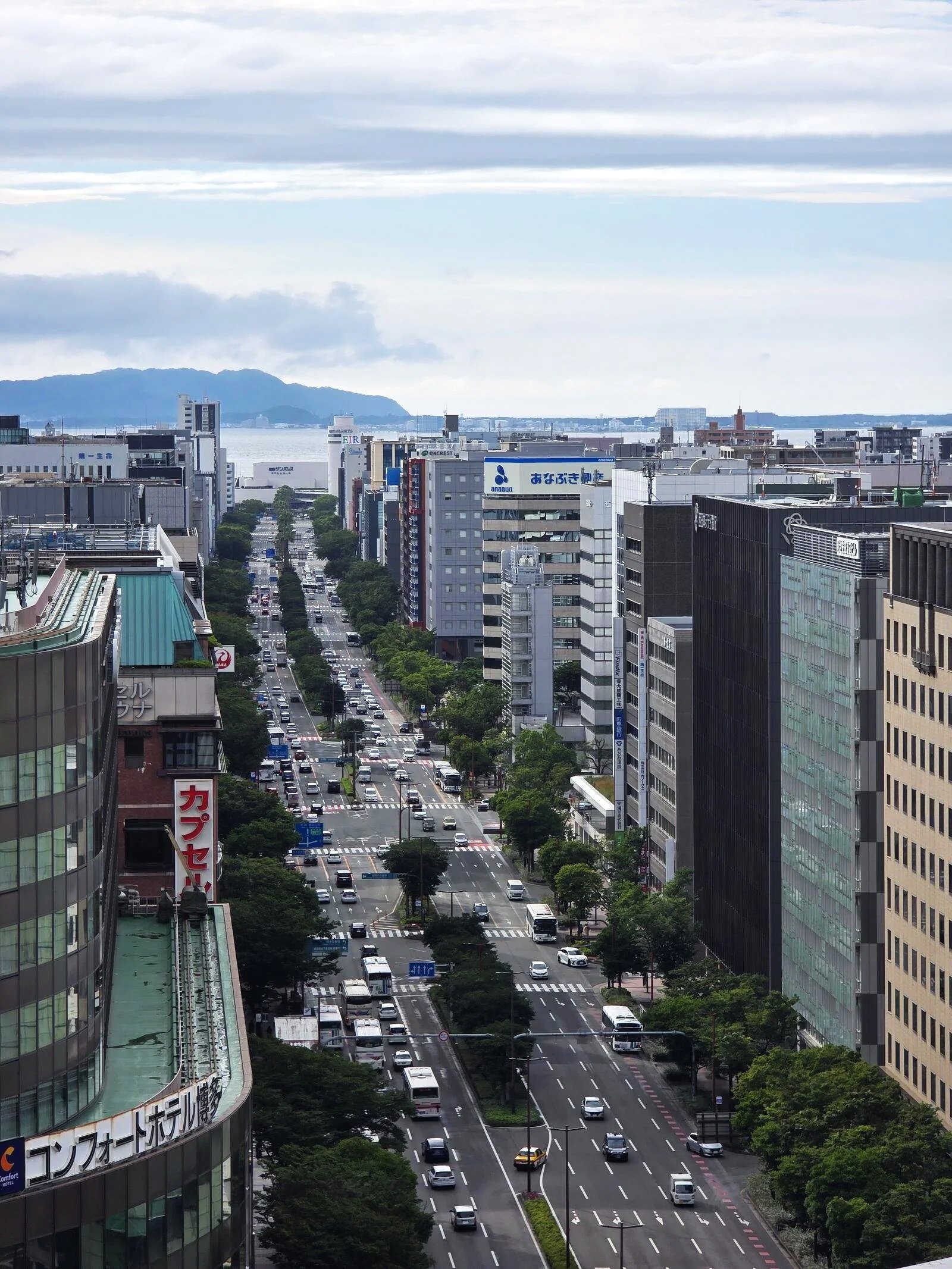 A rooftop view of a city with a large 6-lane road running between tall buildings and the sea and islands visible in the distance