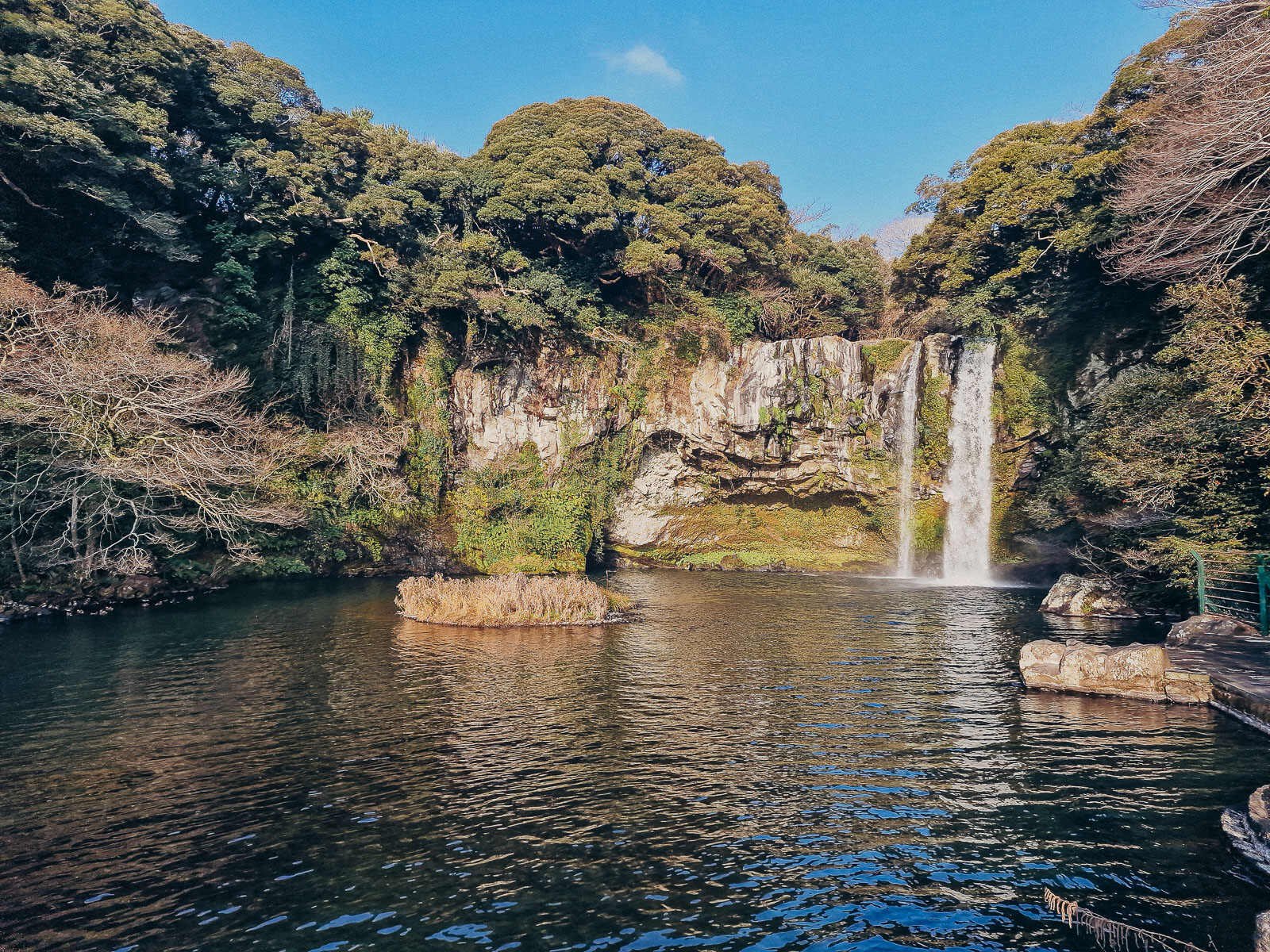 a large pool of water with a waterfall flowing down a cliff face into the pool on the righthand side. The cliffs and pool are surrounded by green trees on a sunny day