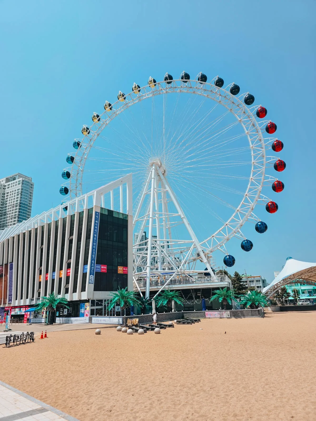 A sandy beach with a white ferris wheel next to a modern balck and white building on a sunny day