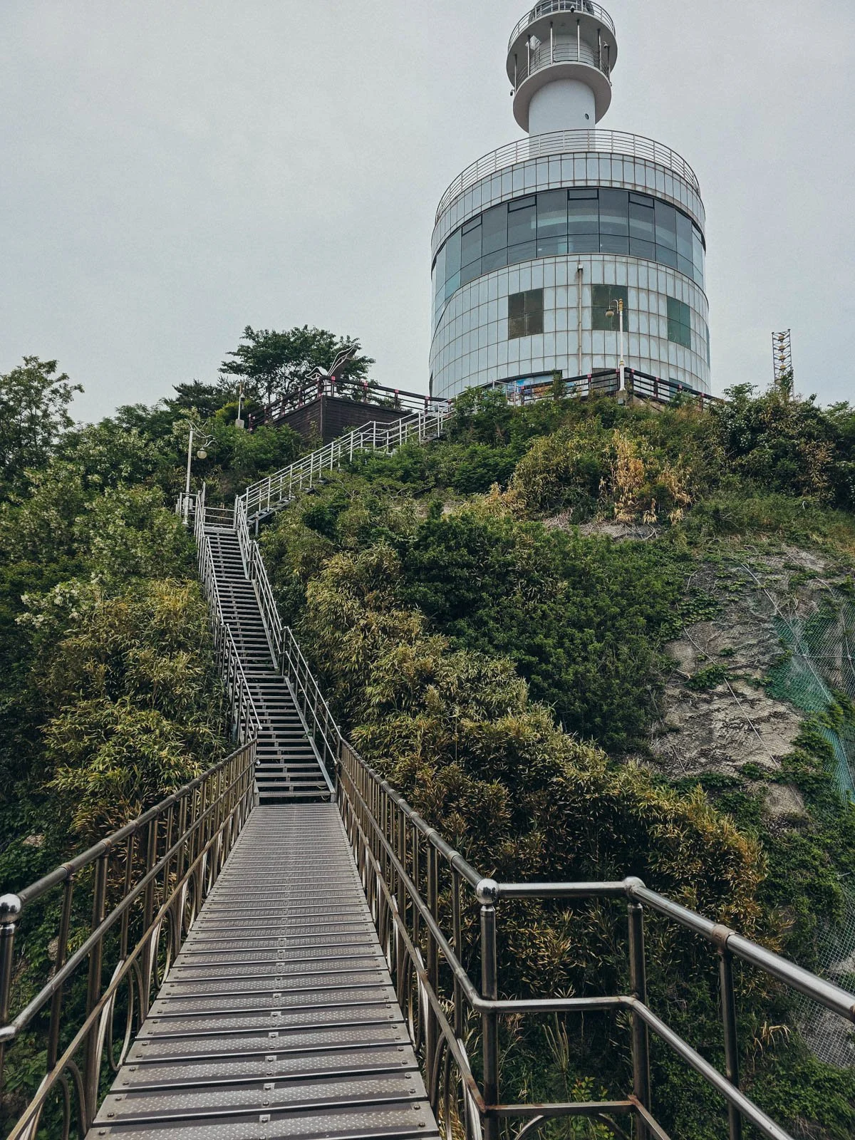 A brutalist style glass lighthouse at the top of a cliff with  steps leading up the side of the cliff which is covered in greenery.