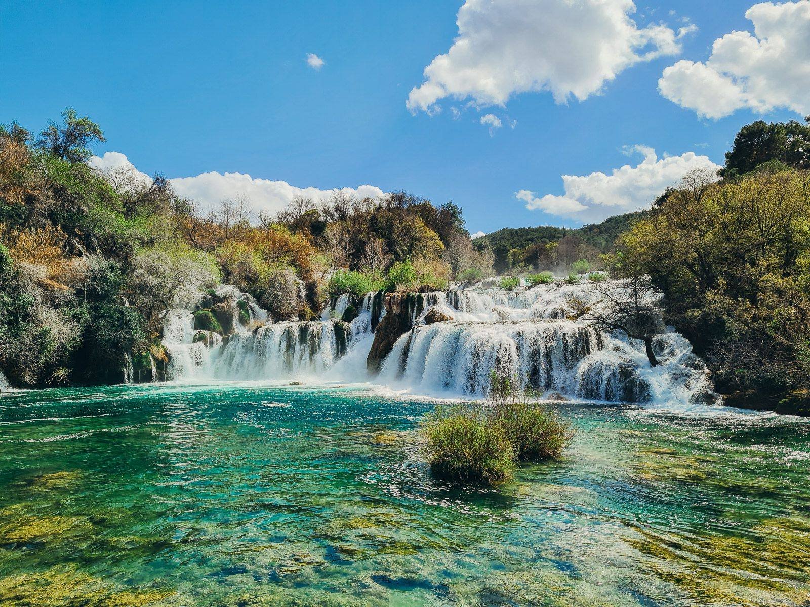 A series of waterfalls flowing white into an emerald green pool. Greenery surrounds the falls and river pool at Krka waterfalls