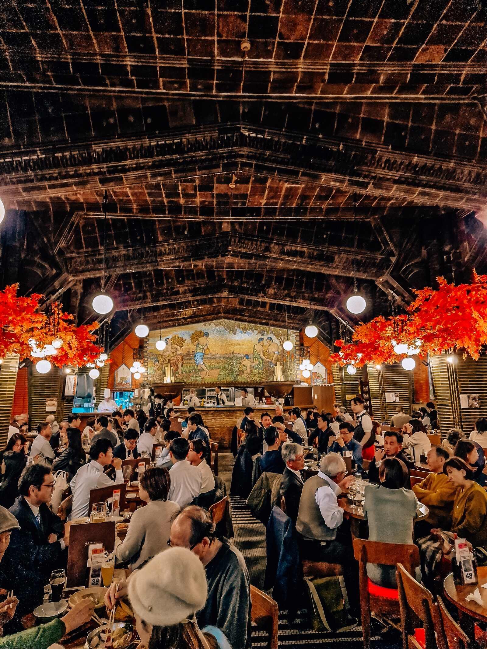 a busy beer hall full of people sitting at tables, the roof is wooden and there's a large mural on the far wall