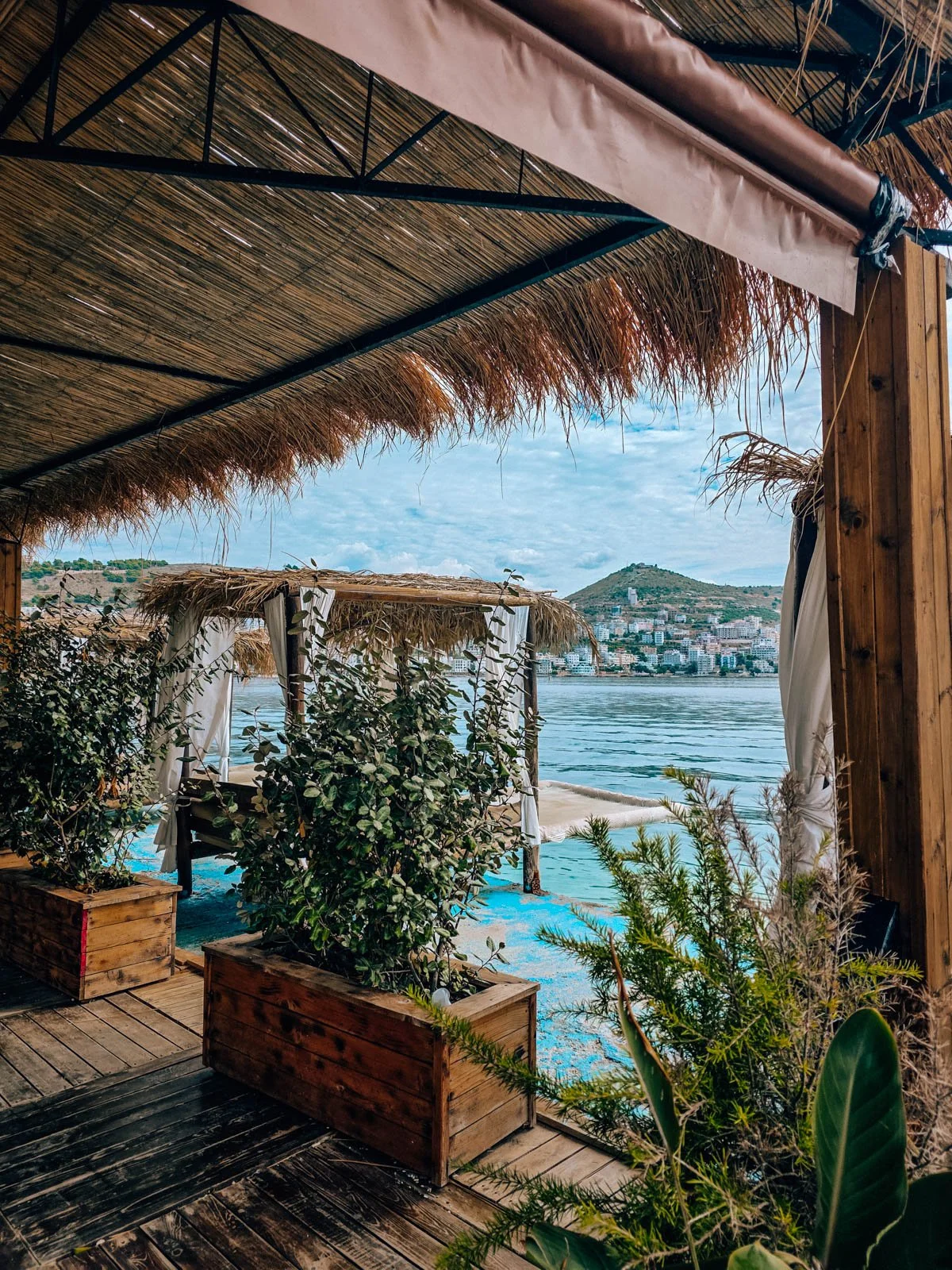 A seaside bar with a wooden deck, many green plants in wooden plant boxes, and a palm frond roof. The bar is along clear blue water with a costal town in the distance