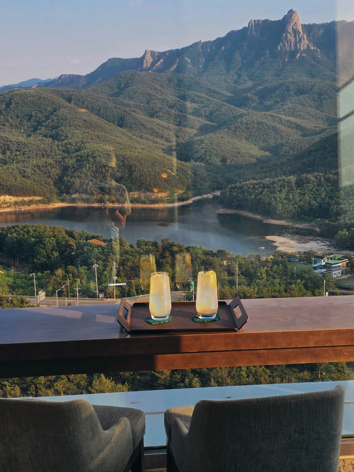Two cold drinks in glasses on a tray on a wooden table. The table is in front of a floor-to-ceiling window that shows a view of rolling tree-covered hills and a lake