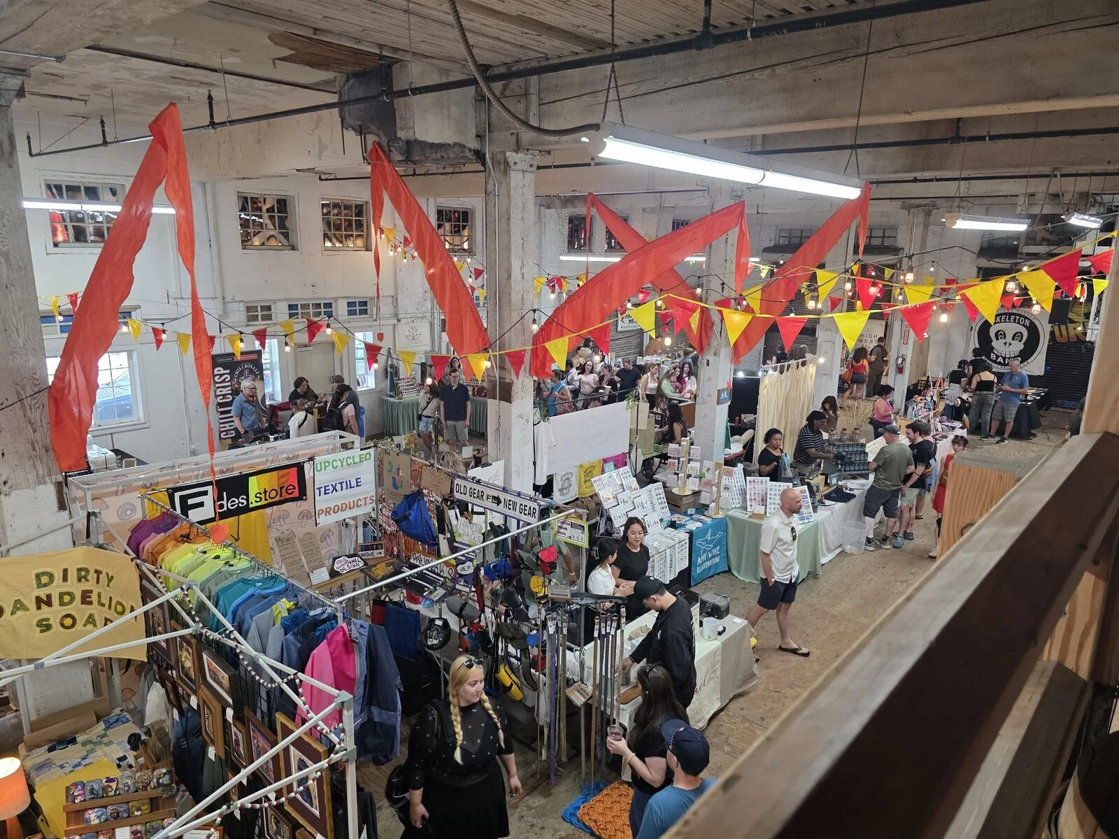 Looking down from a balcony onto an indoor night market in Portland with many stalls selling clothing, food, trickets and red and yellow bunting strung across the large room